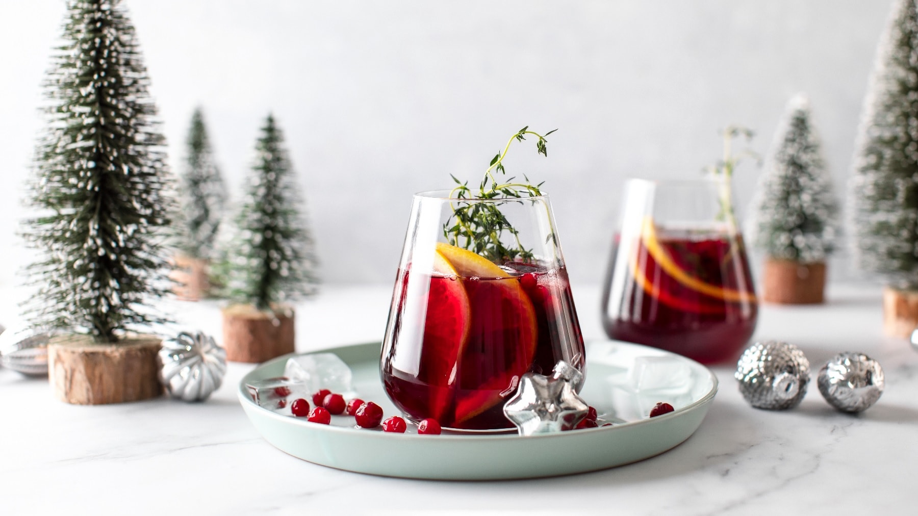 Zwei Gläser vom weihnachtlichen Gin Tonic vor einem weihnachtlichen Hintergrund mit Tannen, silbernen Kugeln und Schnee.