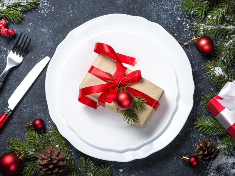 Ein weißer Teller mit einem verpackten Geschenk auf einem grauen und weihnachtlich dekorierten Tisch.