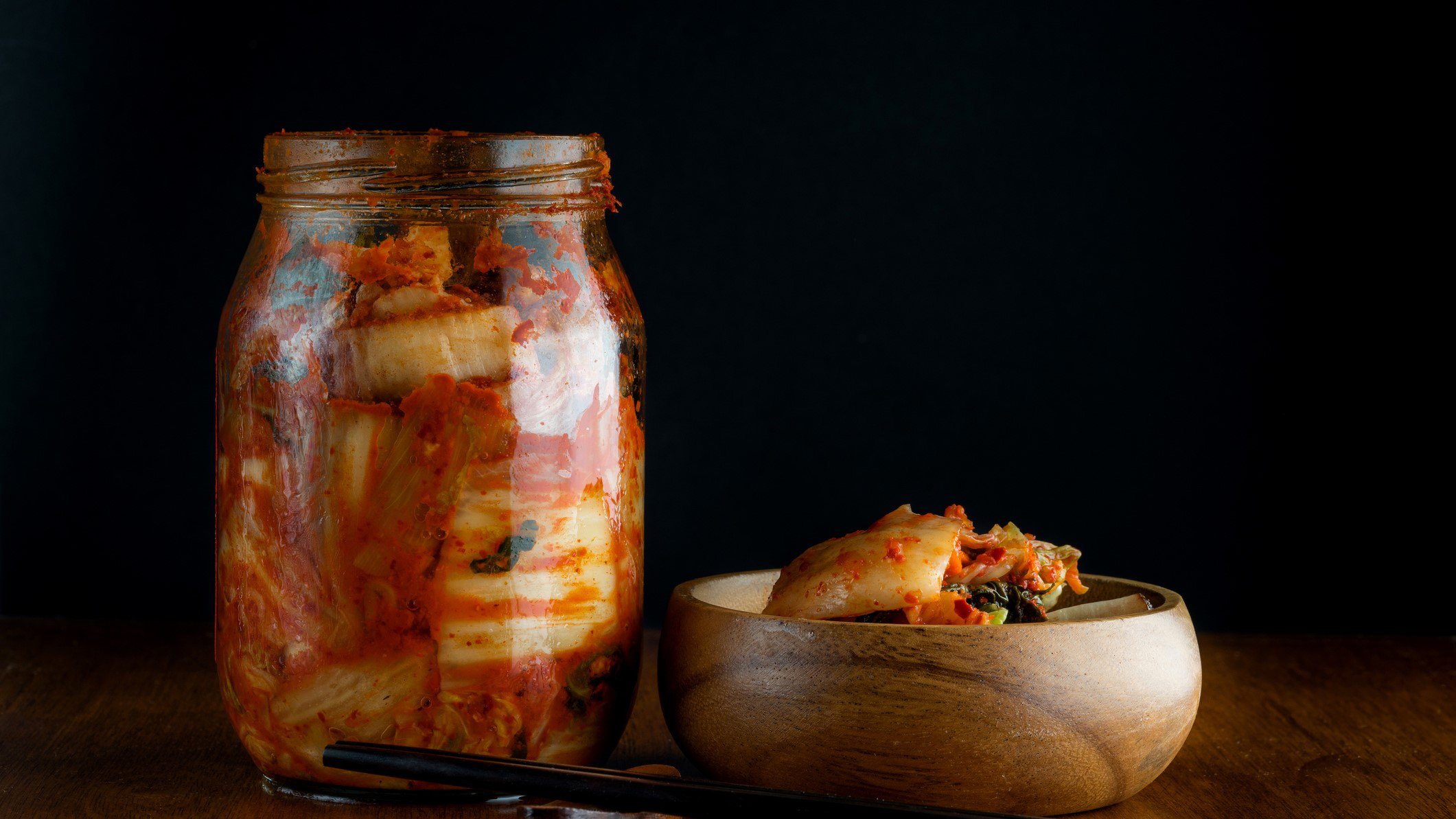 Wirsing haltbar machen mit Wirsing-Kimchi im Glas. Daneben Holzschüssel mit Wirsing-Kimchi und Stäbchen. Frontalansicht.