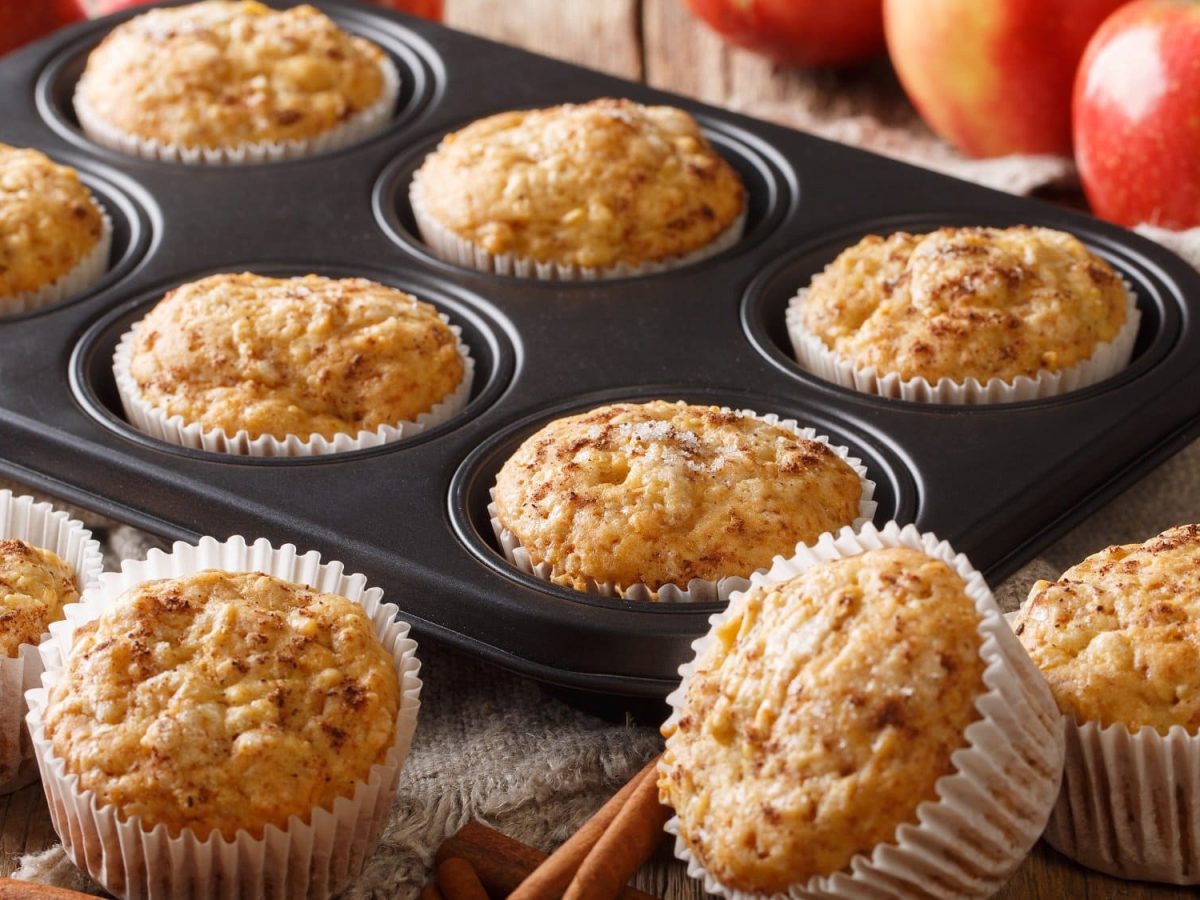 Ein Blech Apfelmus-Muffins, daneben ein paar weitere Muffins sowie Äpfel und Zimtstangen.