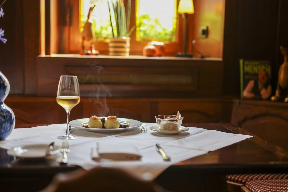 Falstaff Restaurantguide Deutschland: Ein gedeckter Tisch in einem Restaurant, auf dem Tisch ein Glas Weißwein und ein Teller mit dampfenden Klößen.