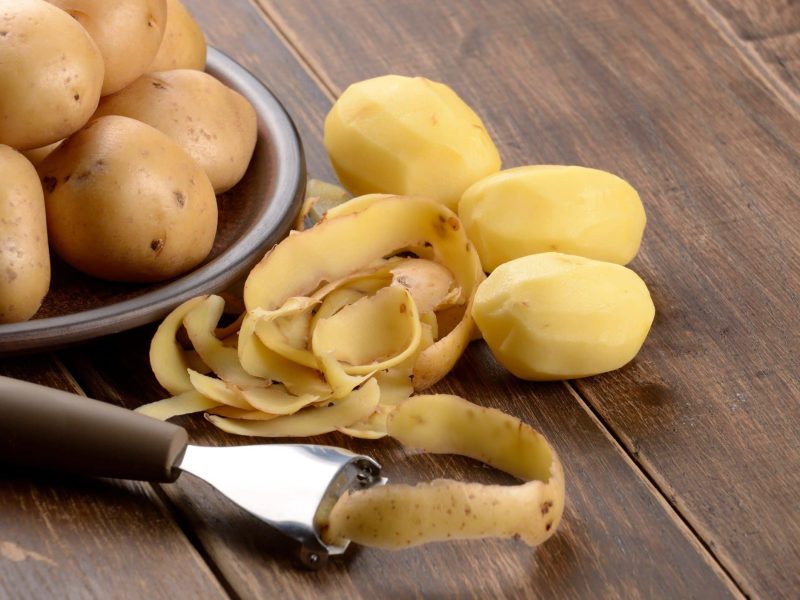 Kann man geschälte Kartoffeln aufbewahren? Ungeschälte Kartoffeln auf einem Teller. Davor geschälte Kartoffeln, Kartoffelschalen und Kartoffelschäler auf einem Holztisch. Draufsicht.