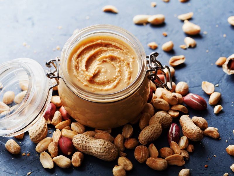 Ist Erdnussbutter vegan:: Ein Weckglas Erdnussbutter, drumherum ganze und geschälte Erdnüsse.