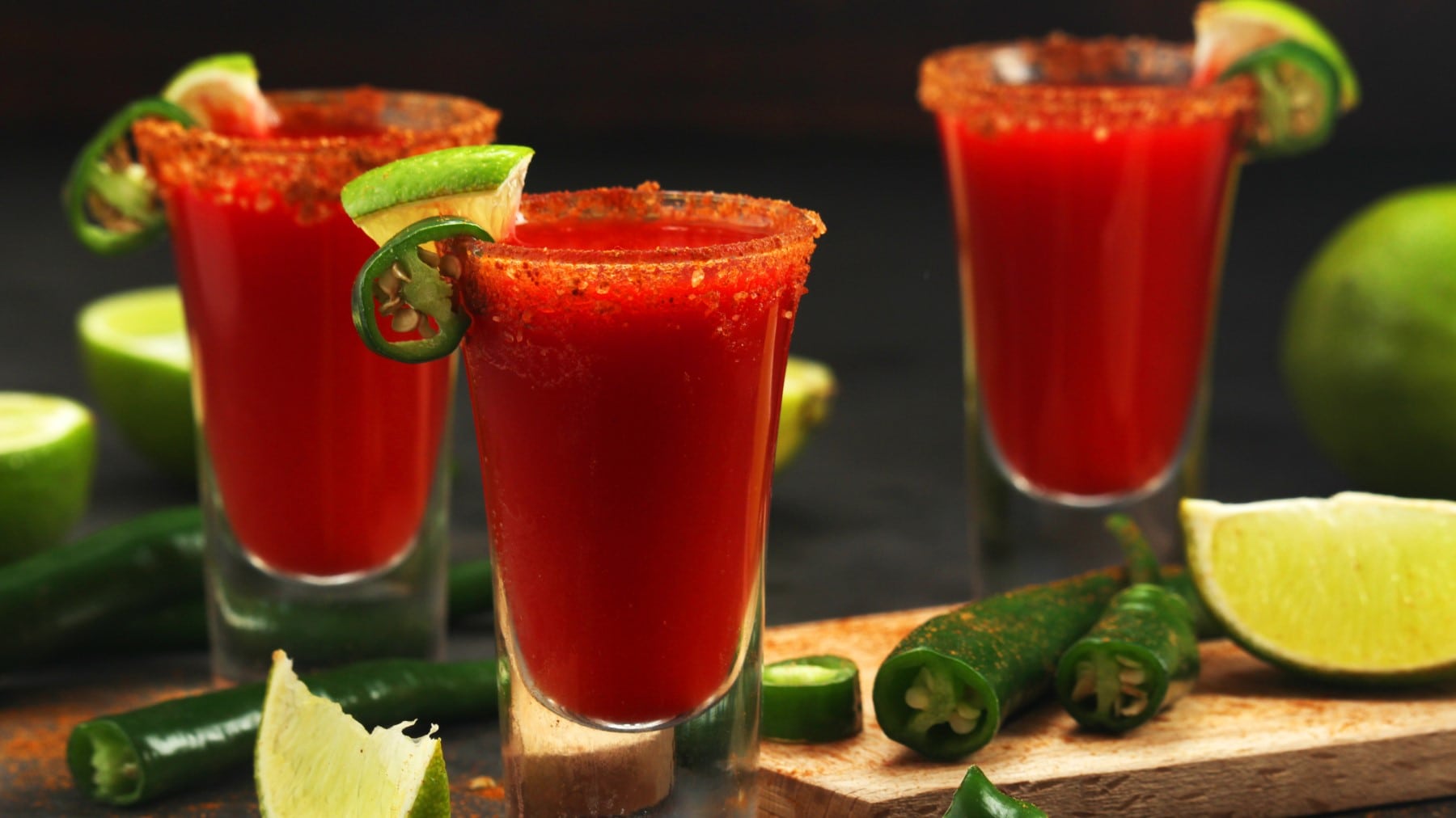 Drei Shotgläser mit Mexikaner Shot, drum herum Limetten und grüne Chilis vor dunklem Hintergrund.