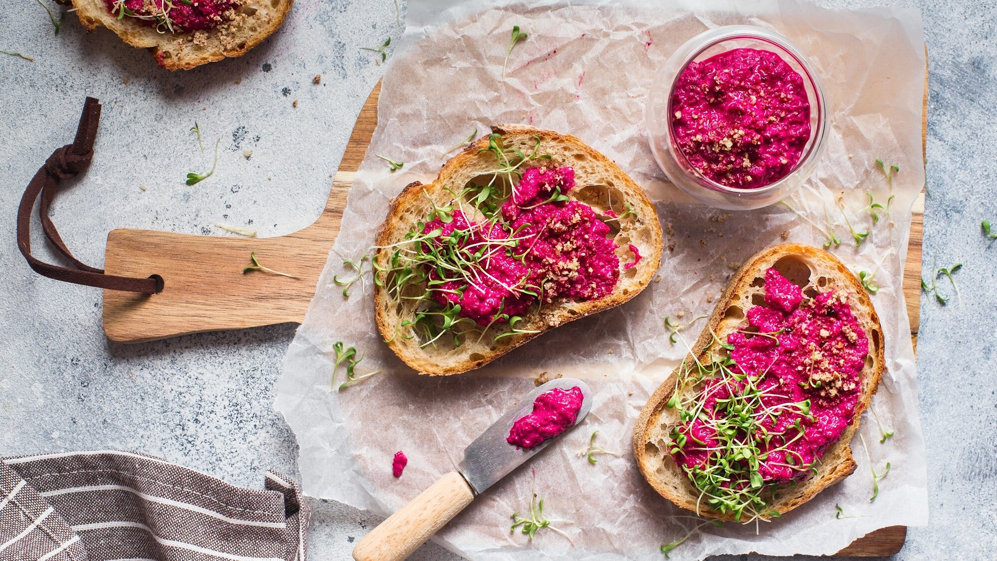 Brote mit Pink Hummus und Kresse auf einem Brett, daneben ein Glas Hummus und ein Messer, alles Draufsicht.