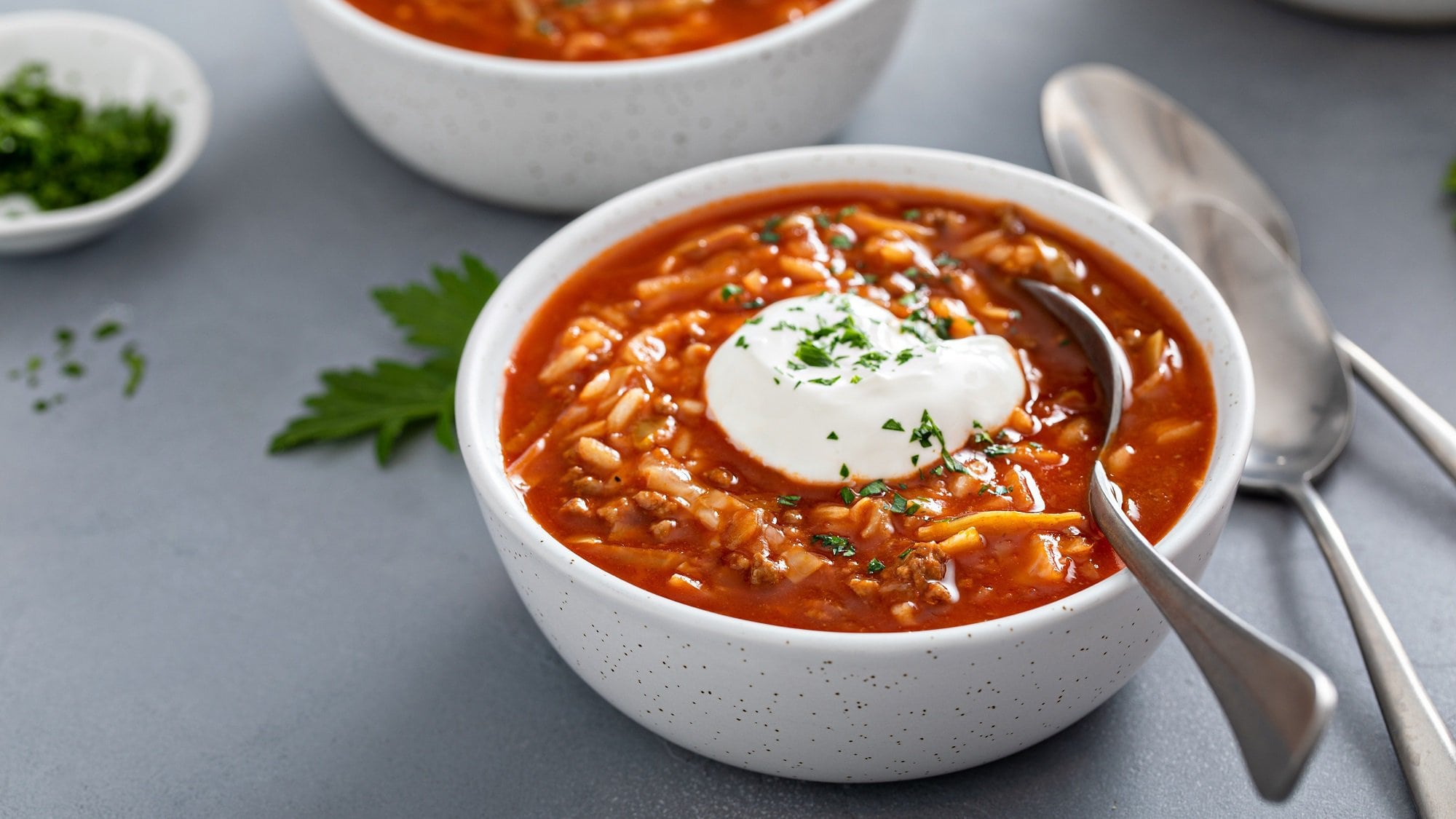 Eine Schüssel Tomaten-Reis-Suppe mit einem Klecks Crème fraîche und einem Löffel darin, daneben liegen weitere Löffel.