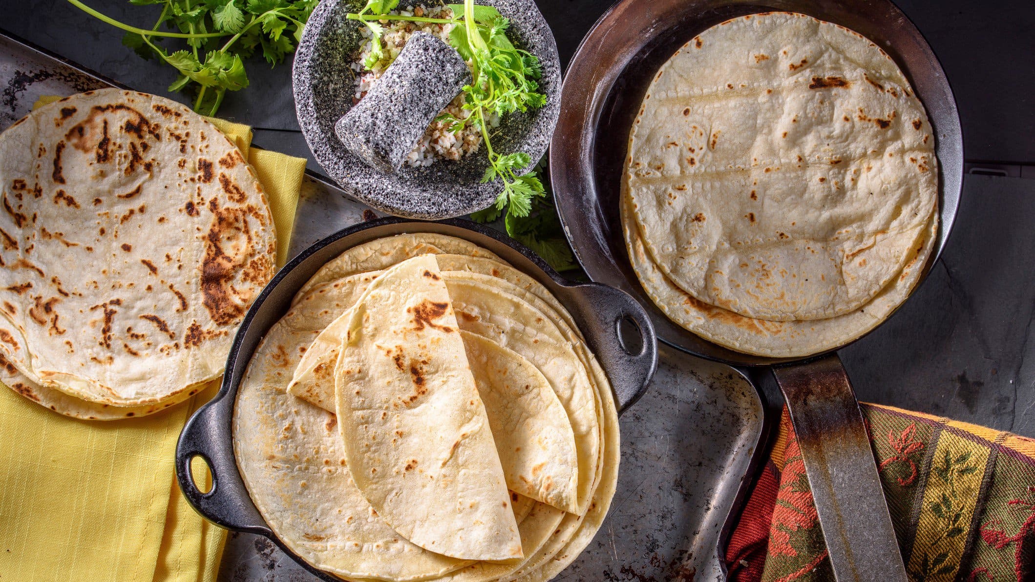 Tortillas aus Maismehl und aus Weizenmehl auf Tellern und in Pfannen verteilt. Im Bild Füllungen und Toppings. Links und rechts Tücher im mexikanischen Stil. Draufsicht.