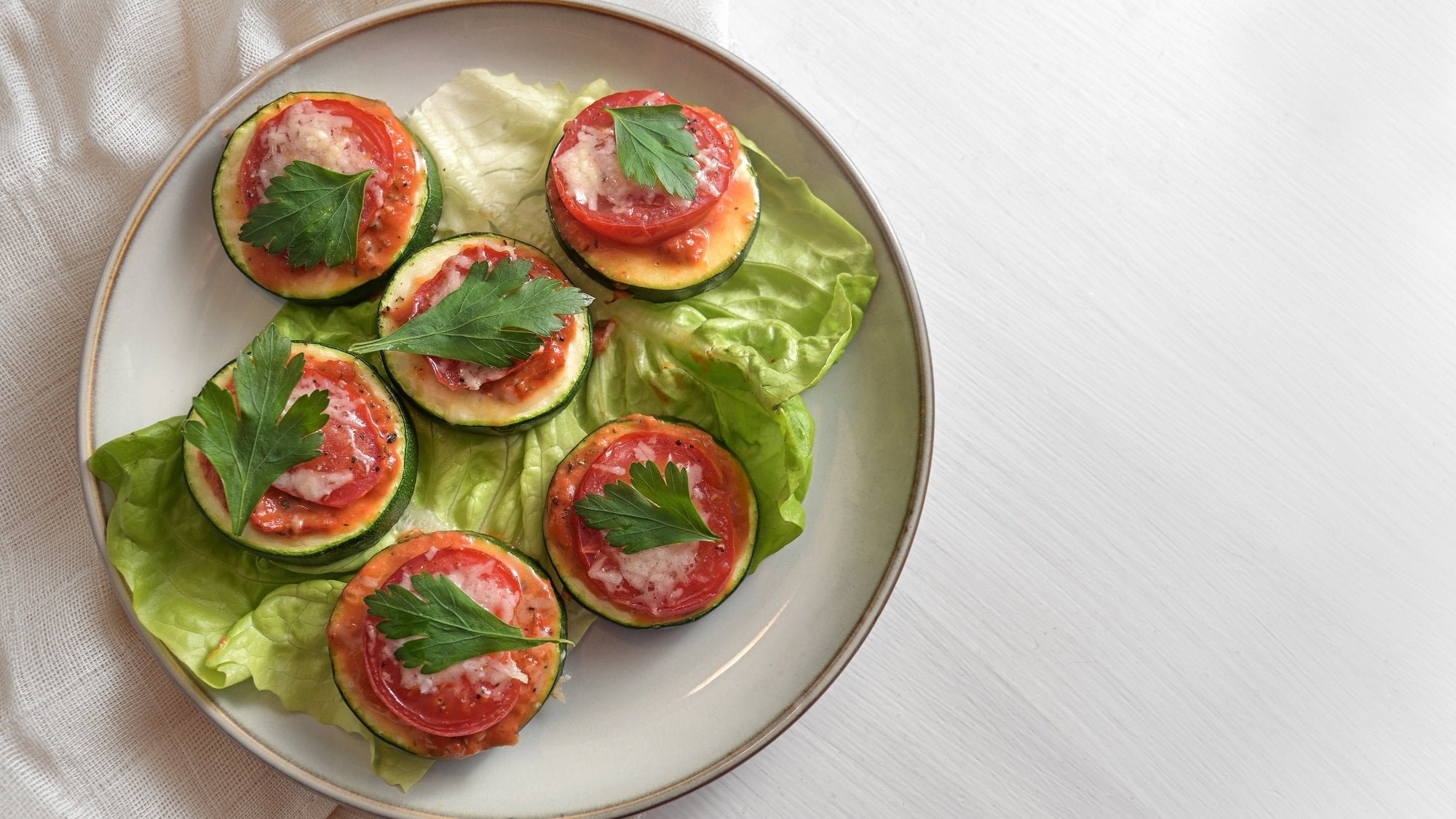 Mehrere Zucchini-Bruschetta-Häppchen mit einer Tomate und Petersilie obenauf auf einem Teller, Draufsicht.