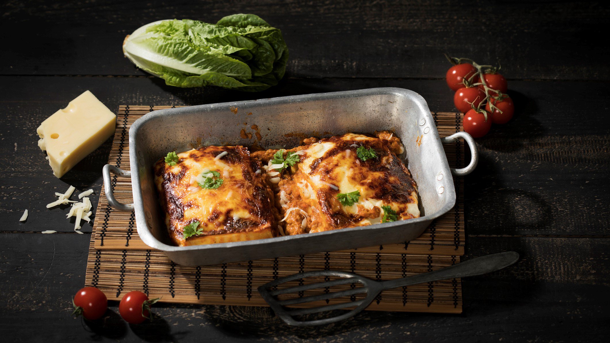 4-Käse-Lasagne mit Knoblauch-Ricottacreme und Parmesan -Crumble in Kasserole auf Bast auf dunklem Untergrund. Im Bild ein Stück Parmesan, einige Tomaten und Salatherz. Draufsicht.