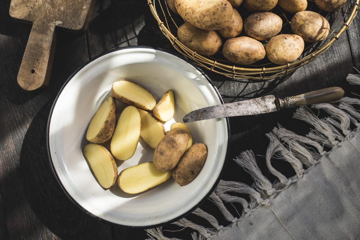 Eine weiße Schüssel mit halbierten Kartoffeln auf einem dunklen Tisch. Daneben ein Korb mit Kartoffeln und eine graue Decke.