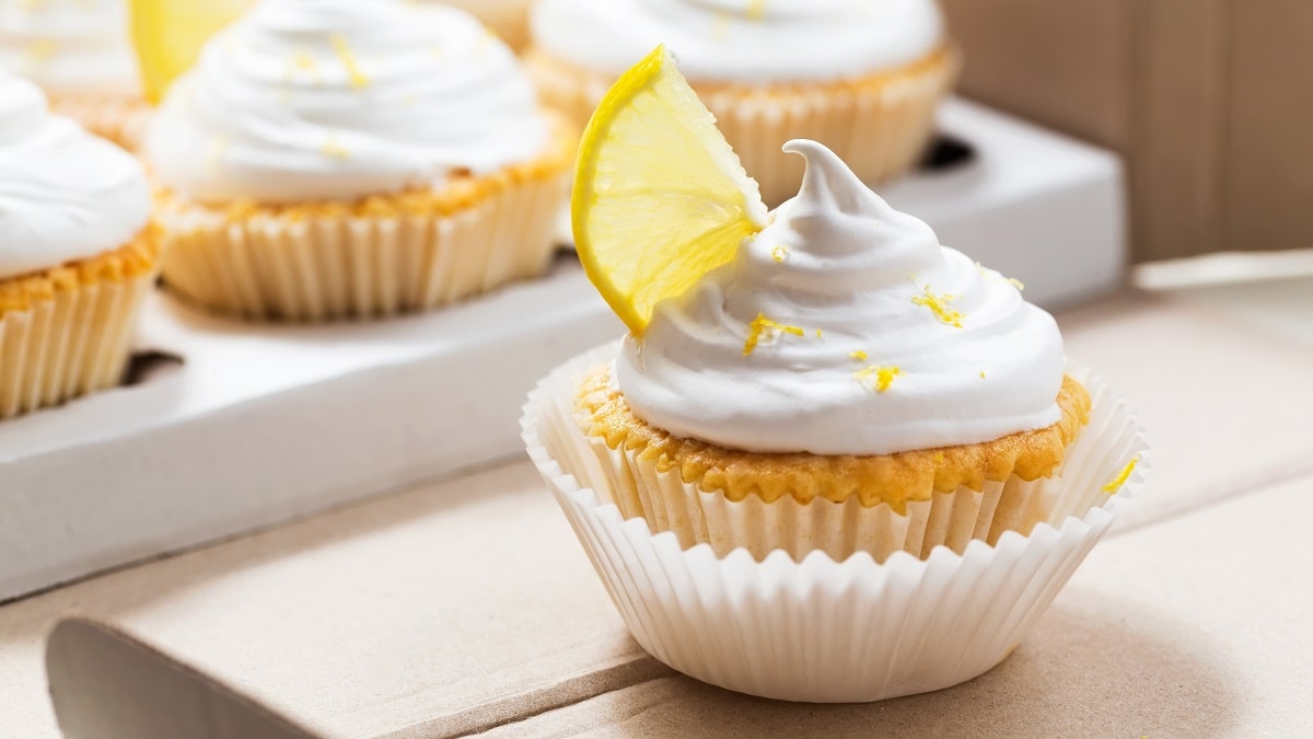 Frontalsicht. Mehrere Lemon-Cupcakes stehen auf einem Untergrund aus Holt. Sie haben eine weiße Haue in dem ein Zitronenschnitzer steckt-