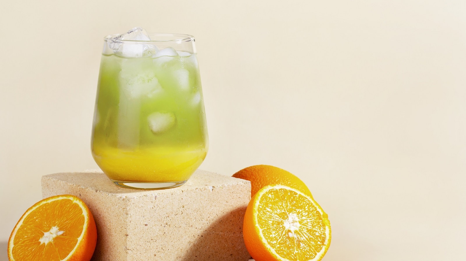 Ein Glas mit Matcha-Limonade auf einem beigen Ziegelstein neben frischen Orangen.