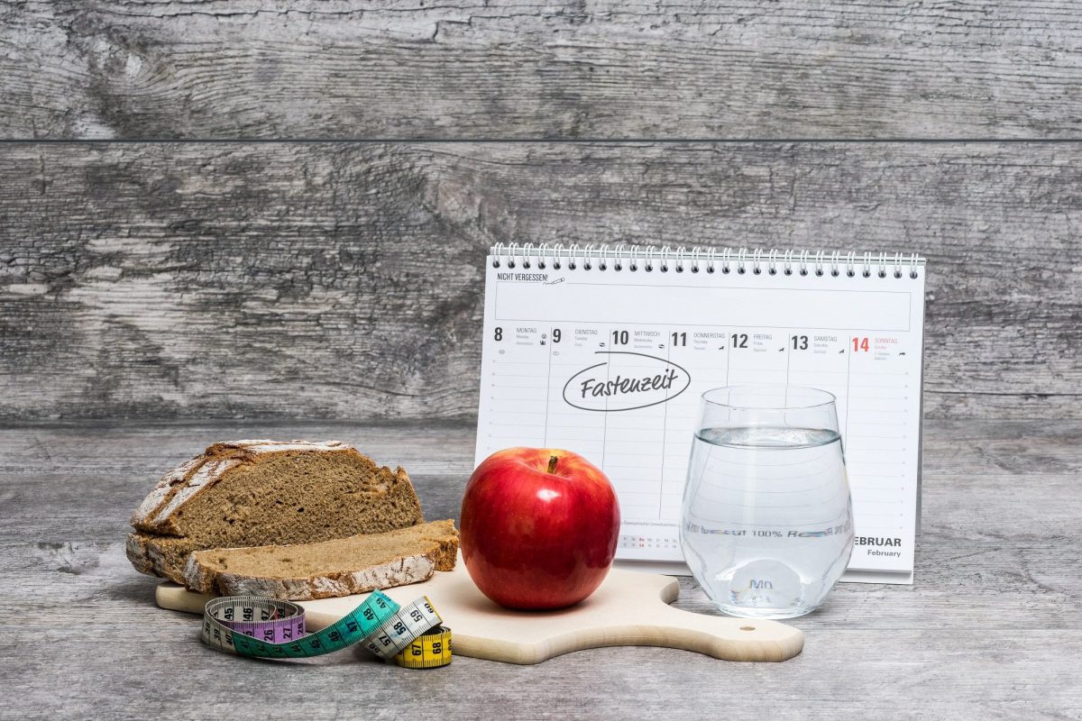 Fasten nach Karneval: Ein Kalender, in dem die Fastenzeit eingetragen ist, davor ein Apfel, ein Glas Wasser, Brot und ein Maßband.
