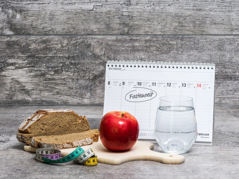 Fasten nach Karneval: Ein Kalender, in dem die Fastenzeit eingetragen ist, davor ein Apfel, ein Glas Wasser, Brot und ein Maßband.