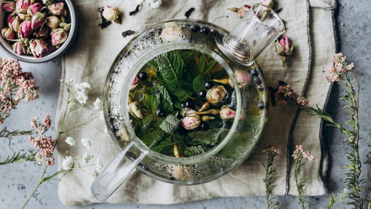 Draufsicht: Grauer Untergrund mit einem Stofftuch auf dem eine Glasteekanne steht in der Kräuter-Rosen-Tee aufgegossen ist. Um die Kanne herum liegen einzelne Blüten und Blätter.