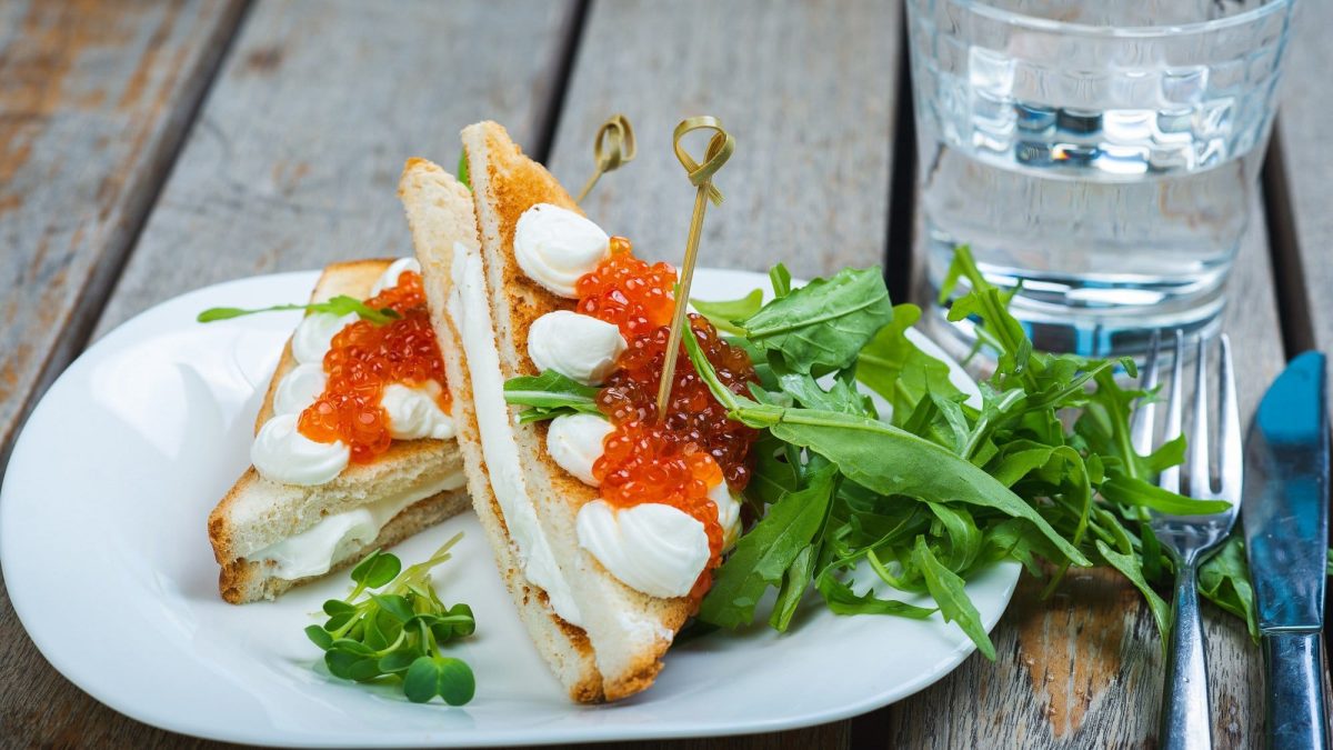 Frontalsicht: Auf einem Holztisch steht einen weißer Teller. Auf dem Teller lieben zwei Mozzarella-Toast mit Forellenkaviar die rechteckig zugeschnitten sind.