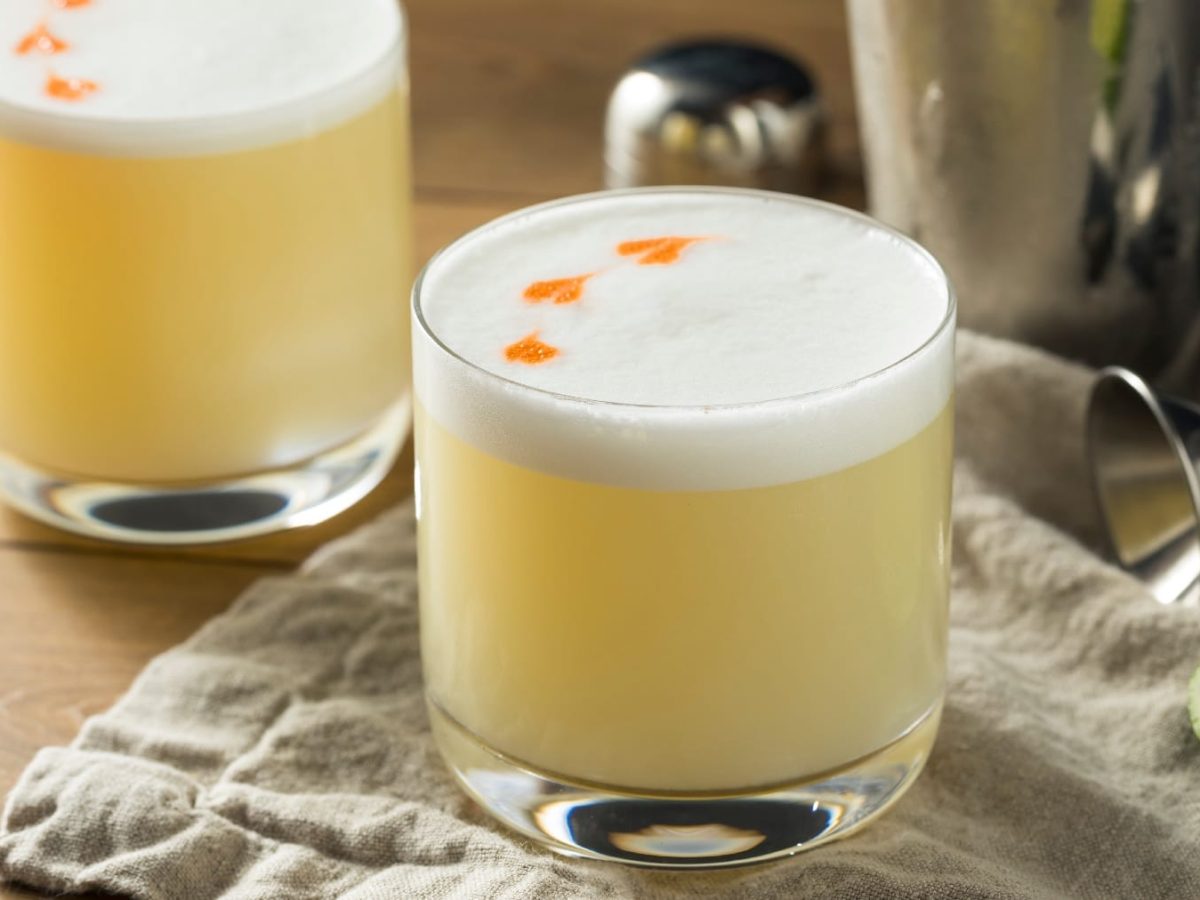 Zwei Gläser mit Pisco Sour auf einem Holztisch mit Limette und siulbernem Cocktail-Shaker.