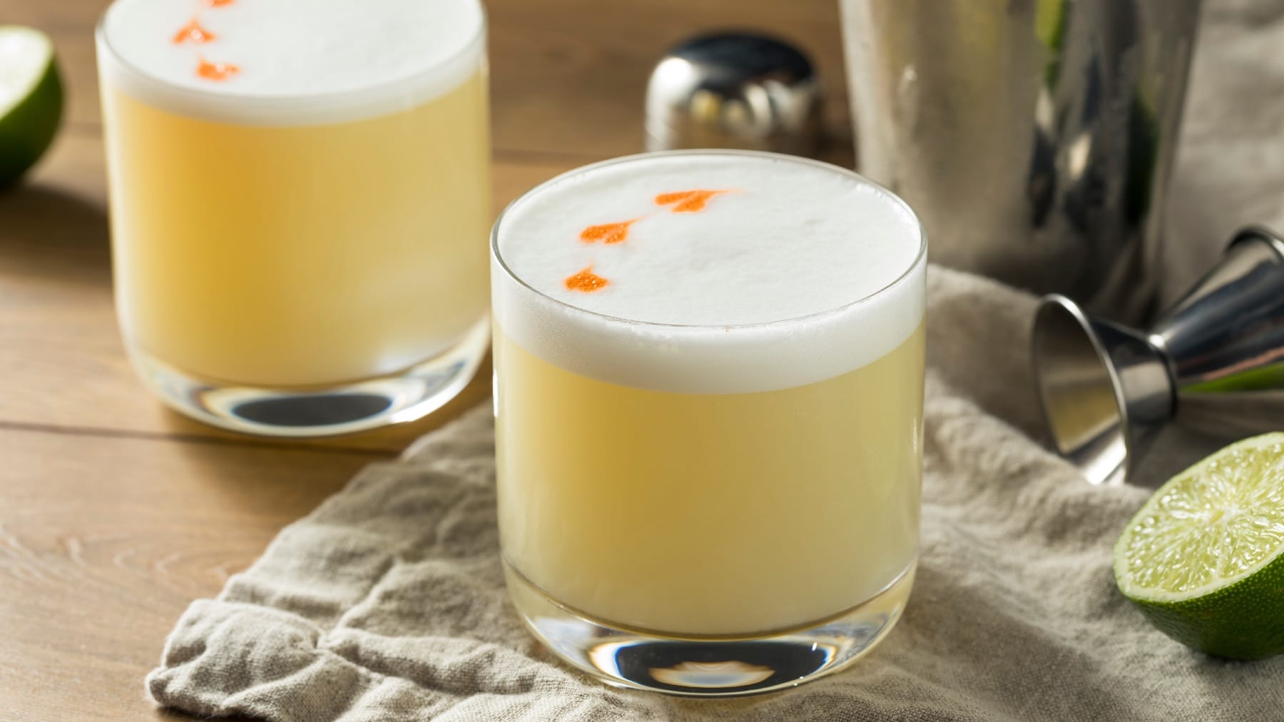 Zwei Gläser mit Pisco Sour auf einem Holztisch mit Limette und siulbernem Cocktail-Shaker.