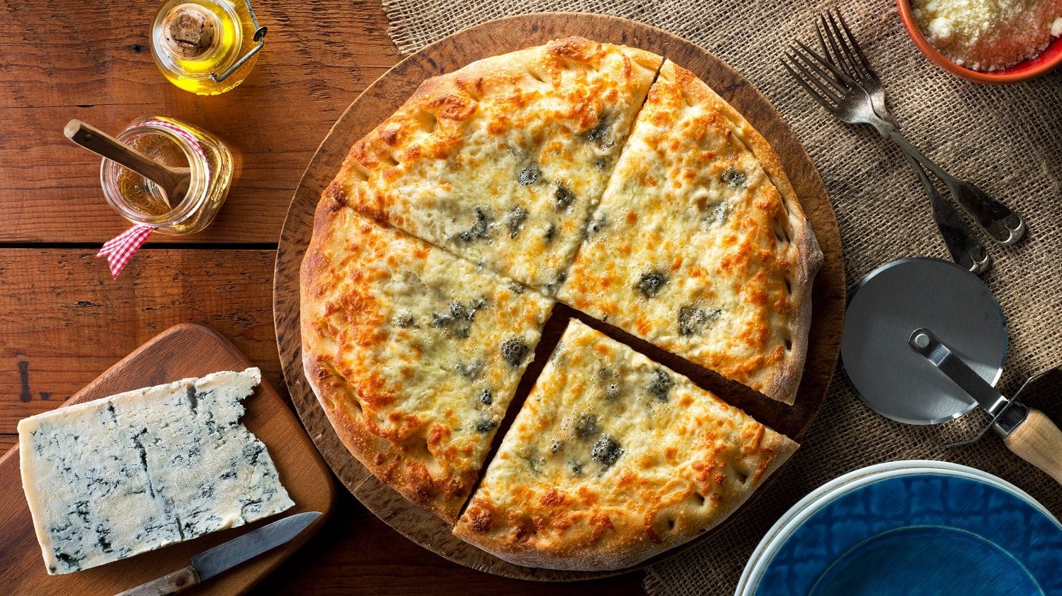 Draufsicht: eine Pizza Margherita mit Blauschimmelkäse die in 4 teile geschnitten wurde liegt auf einem Holztisch. Daneben liegt ein Blauschimmelkäse sowie ein Besteck.