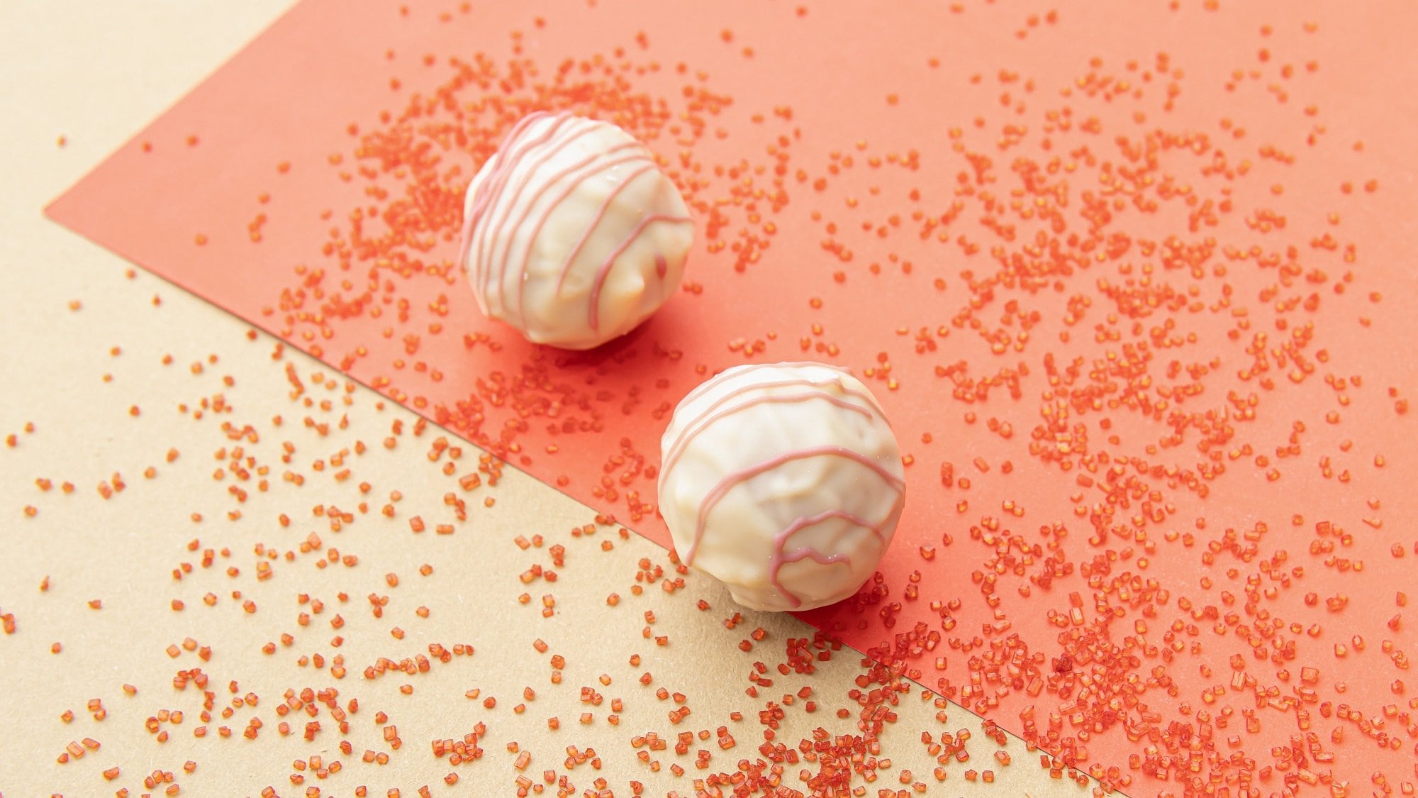 Zwei Red-Velvet-Pralinen mit weißer Schokolade überzogen, drumherum roten Zuckerperlen auf rot-weißem Untergrund.