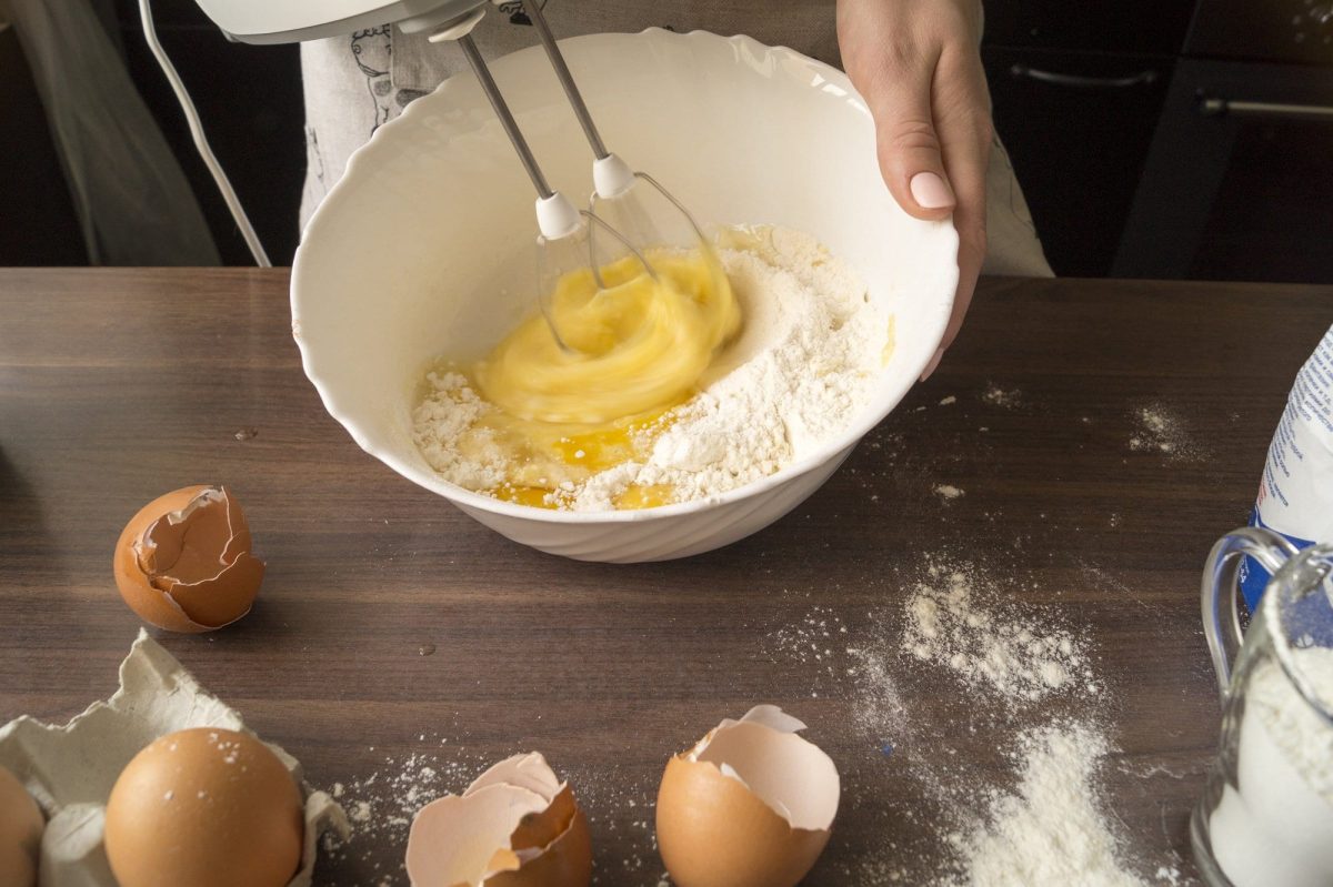 auf einen dunklem Holztisch liegen Mehlreste und aufgeschlagene Eierschalen- Eine hand hält eine weiße Schüssel mit Eiern und hält ein Handrührgerät mit Knethaken in der Hand.