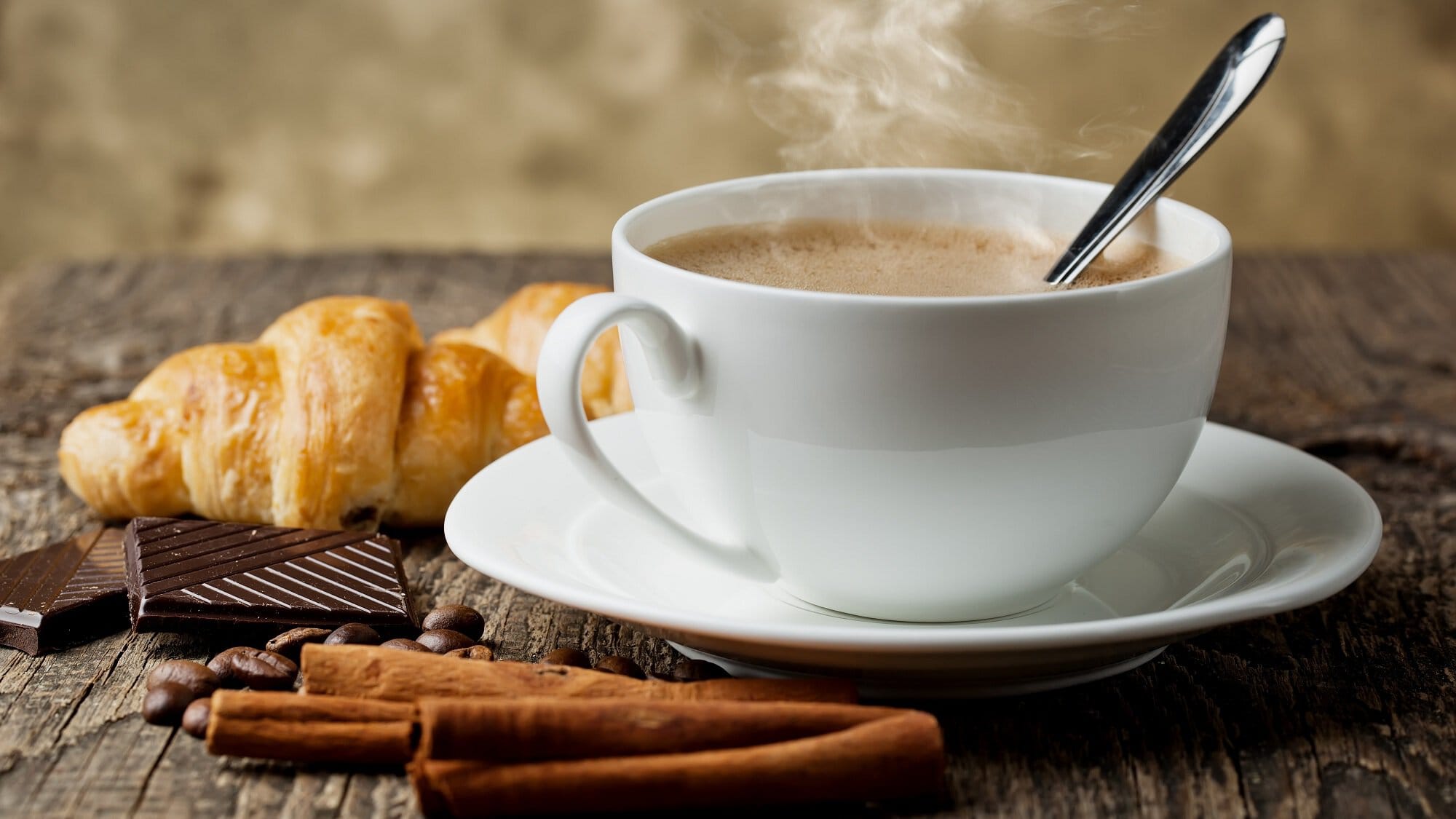Frontalsicht: Ein Kaffee mit Vanille steht auf einem Holztisch. Daneben liegen Vanilleschoten und ein Croissant.