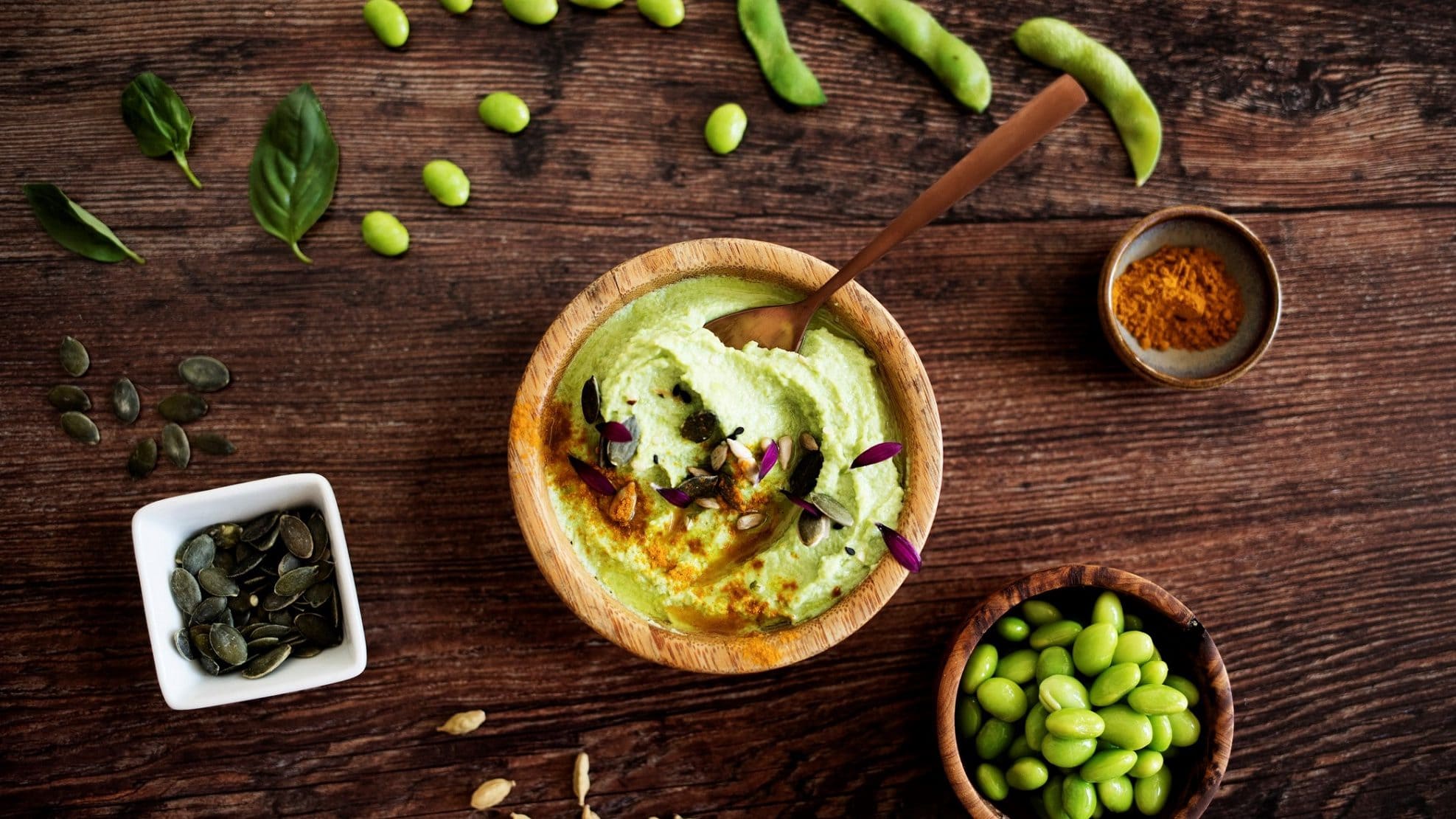Draufsicht: Schüssel Edamame-Hummus mit Kürbis- und Edamame-Bohnen und Currypulver auf einem Holztisch.