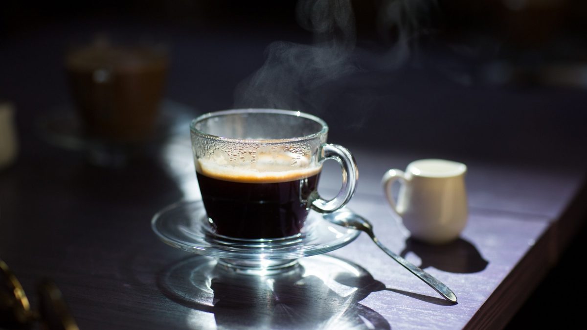 Frontalsicht: Auf auf einem dunklen Tisch stehen vor einem dunklen Hintergrund ein Glas schwarzer Kaffee sowie ein helles weißen Kännchen aus Porzellan.