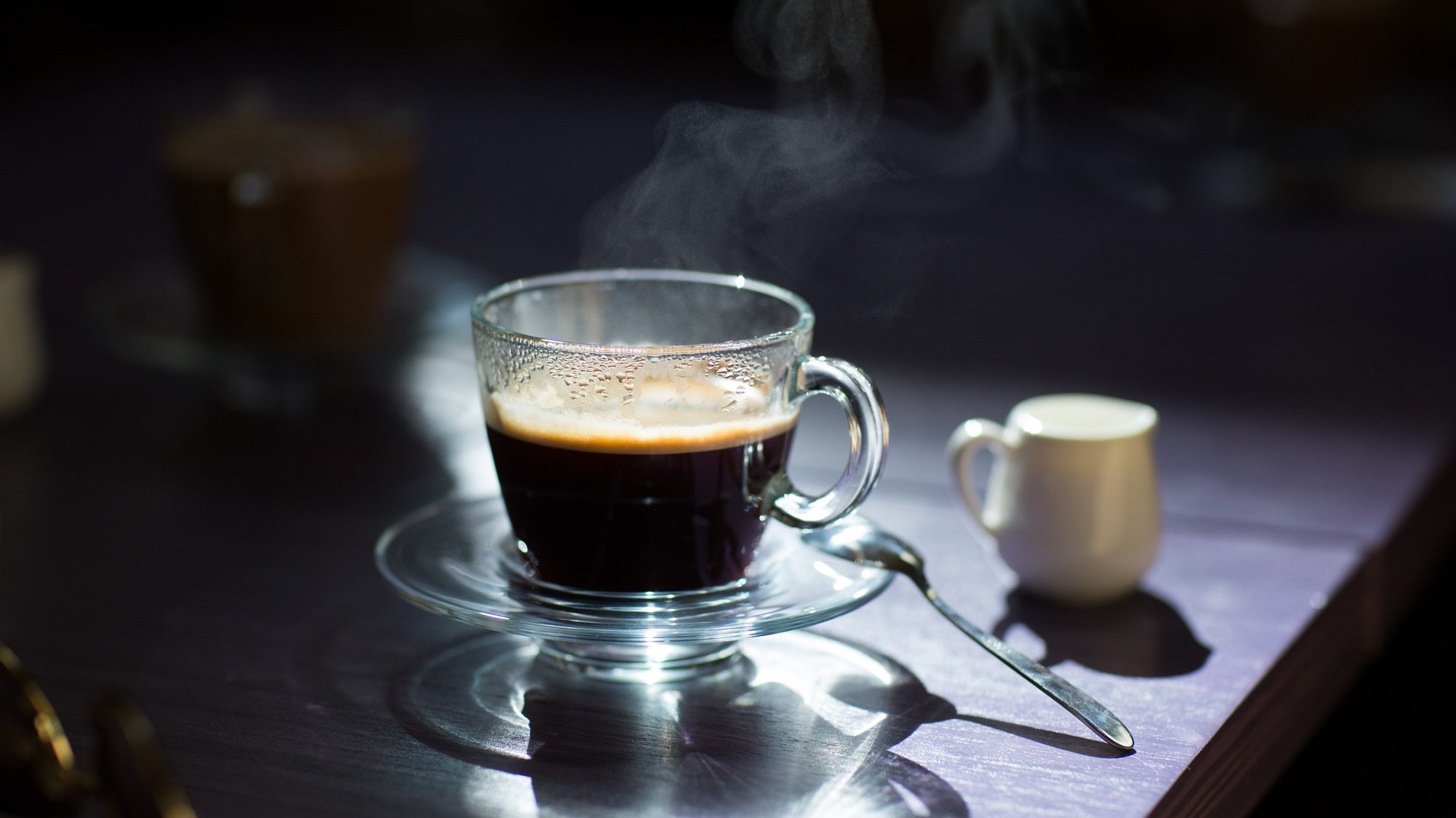 Frontalsicht: Auf auf einem dunklen Tisch stehen vor einem dunklen Hintergrund ein Glas schwarzer Kaffee sowie ein helles weißen Kännchen aus Porzellan.