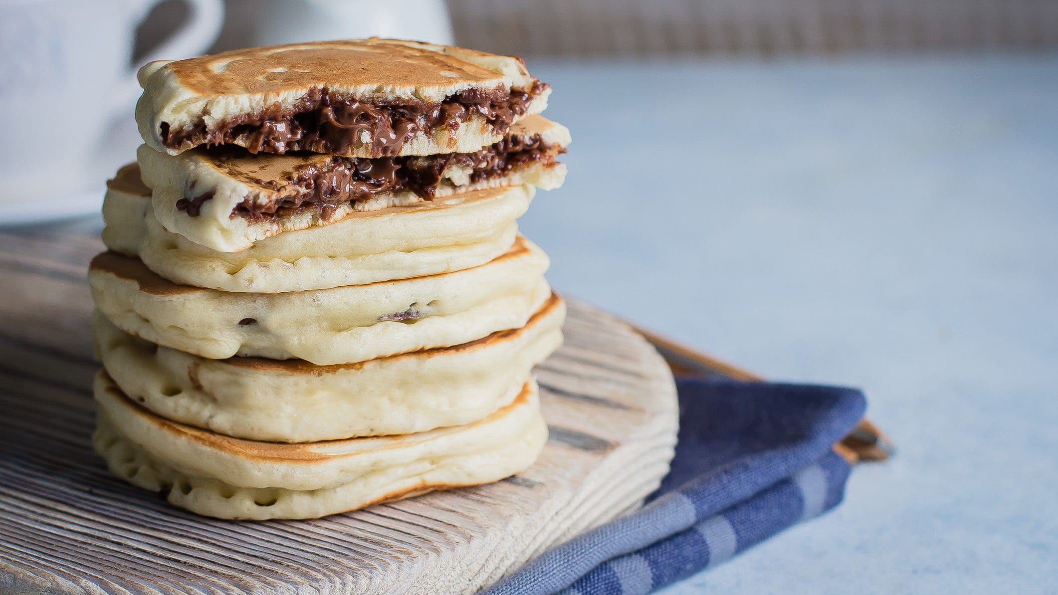 Gefüllte Pancakes mit Nutella gestapelt auf Holzbrett auf Küchentuch. Im Hintergrund eine Tasse. Frontalansicht.