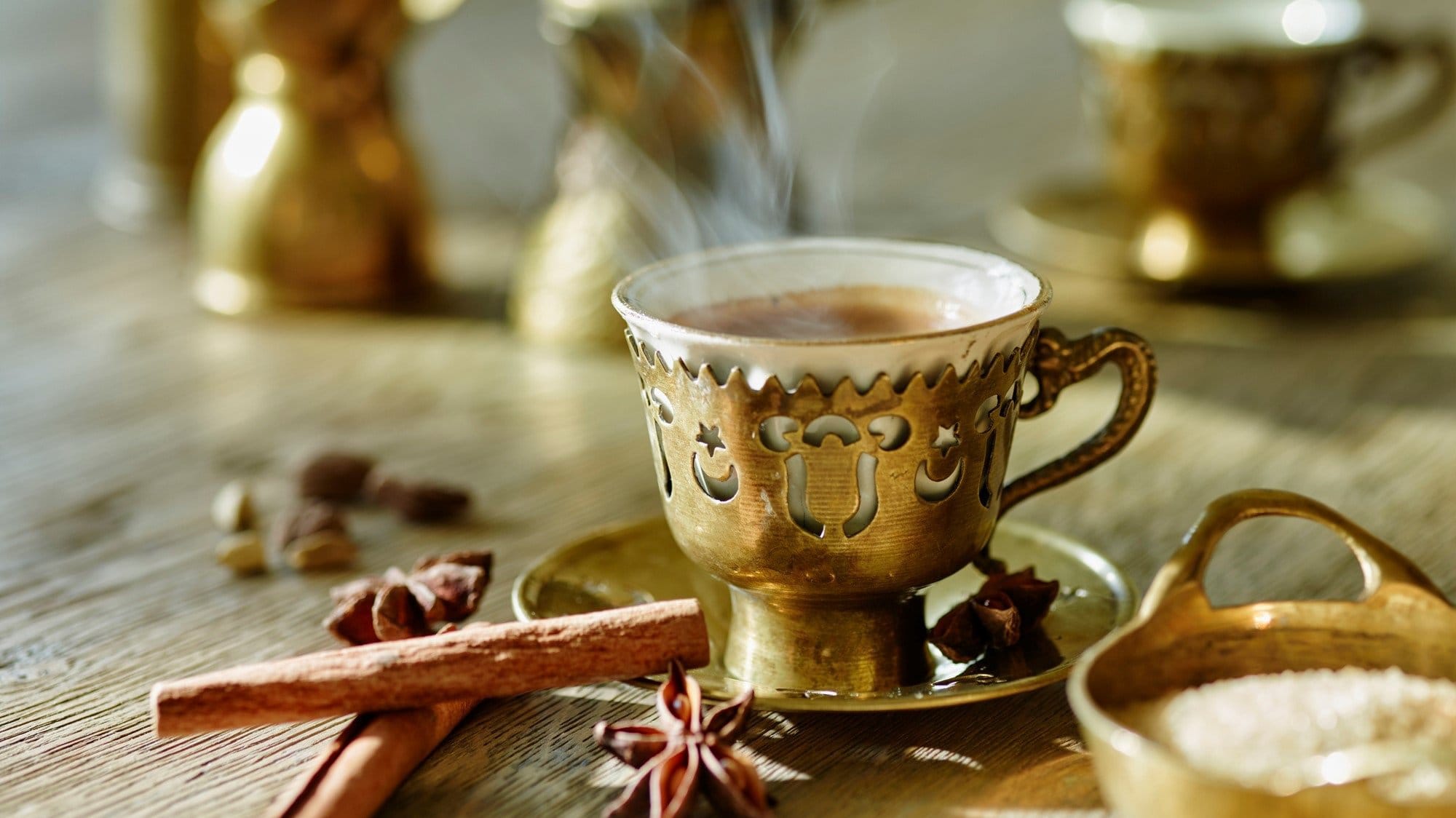 Frontalsicht: Eine arabische Mokkatasse steht auf einem Tisch. Drum herum liegen Gewürze, wie etwa eine Vanilleschote und Sternanis.