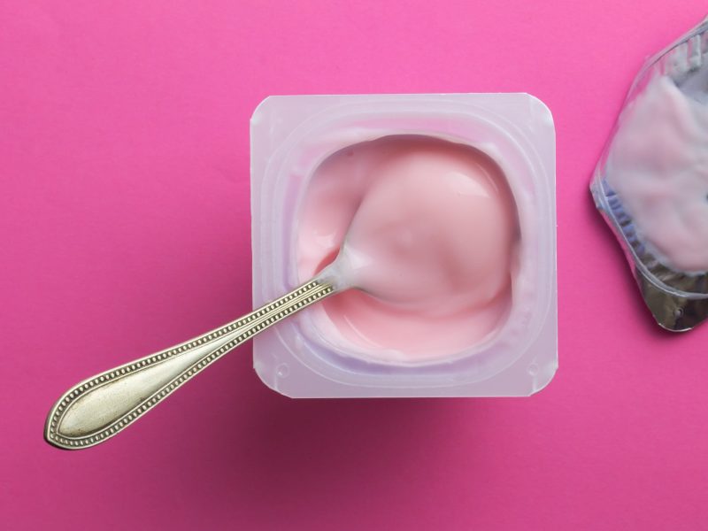 Draufsicht: Auf einem pinken Untergrund steht ein geöffneter Joghurtbecher. In dem Joghurt steckt ein Löffel. Neben dem Joghurt lieget der Joghurtdeckel.