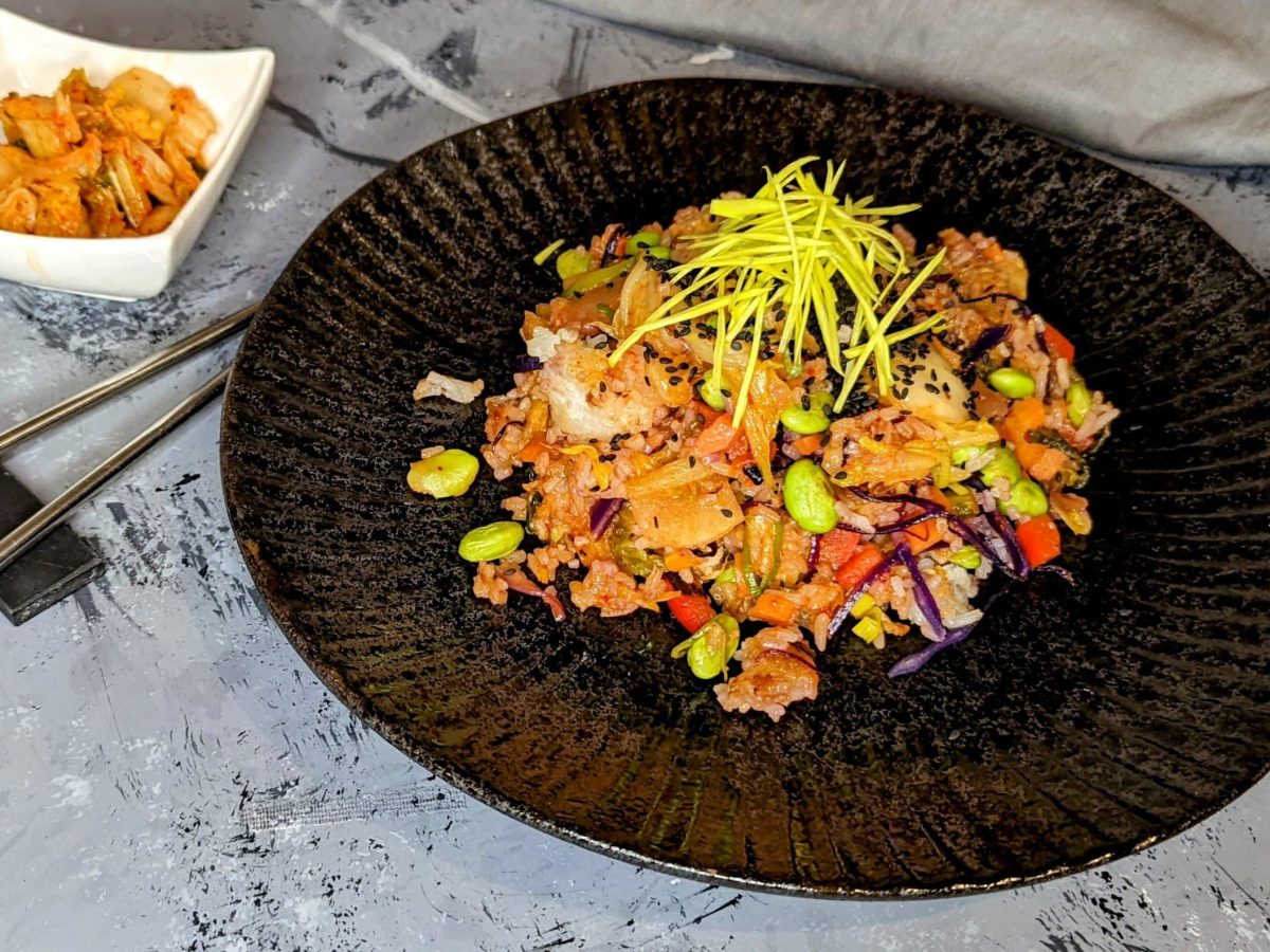 Ein schwarzer Teller mit dem Kimchi-Reis auf einem grauen Untergrund. Daneben Stäbchen und eine kleine weiße Schale mit Kimchi.