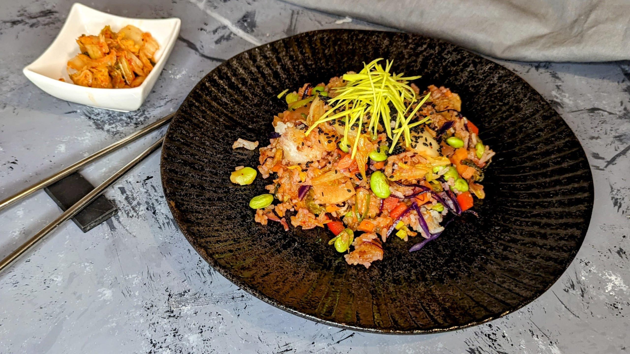 Ein schwarzer Teller mit dem Kimchi-Reis auf einem grauen Untergrund. Daneben Stäbchen und eine kleine weiße Schale mit Kimchi.