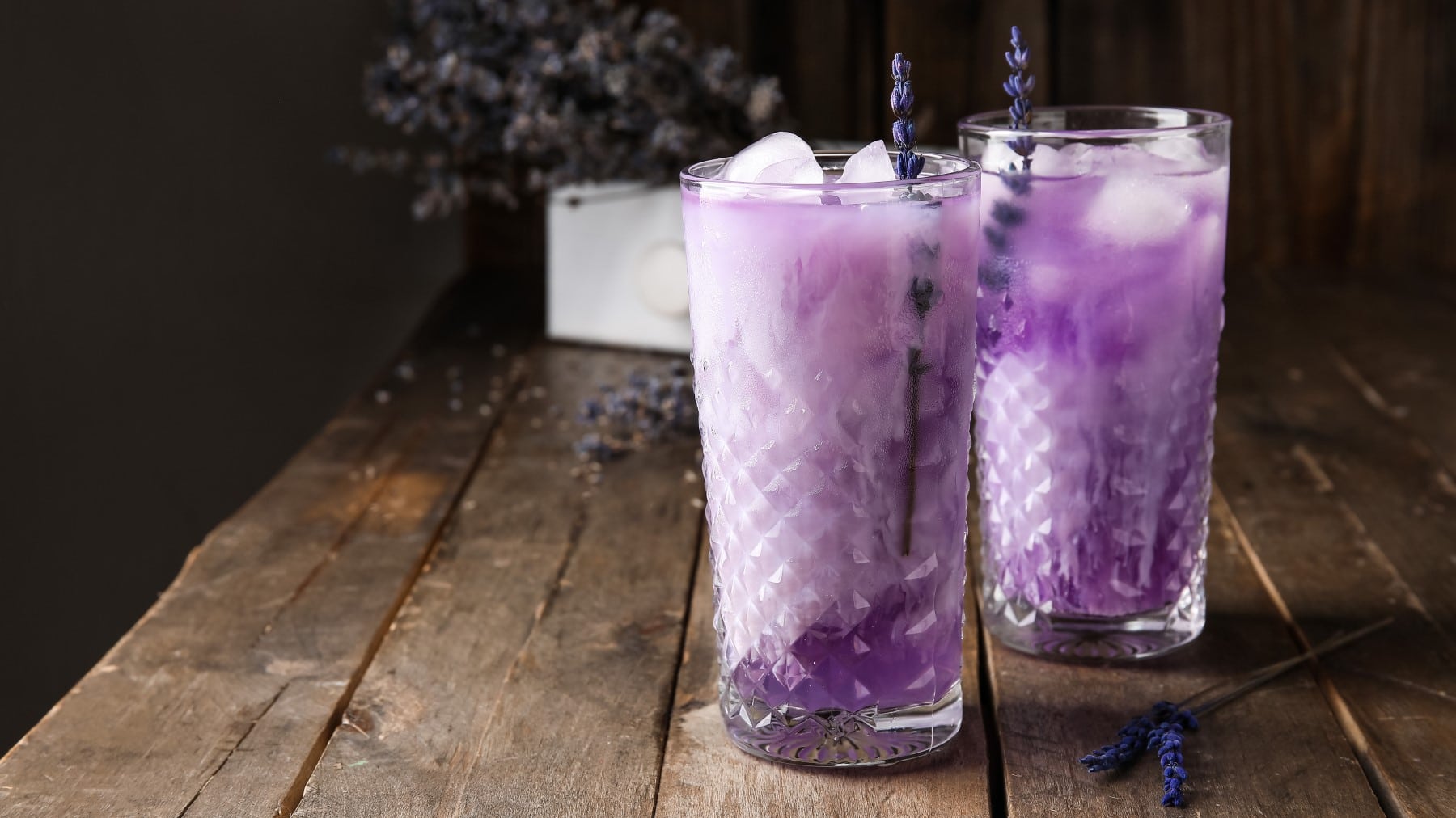 Zwei Gläser mit Lavendel-Cocktail mit Likör 43 mit Eiswürfeln und Strohhalmen auf einem Holztisch vor dunklem Hintergrund.