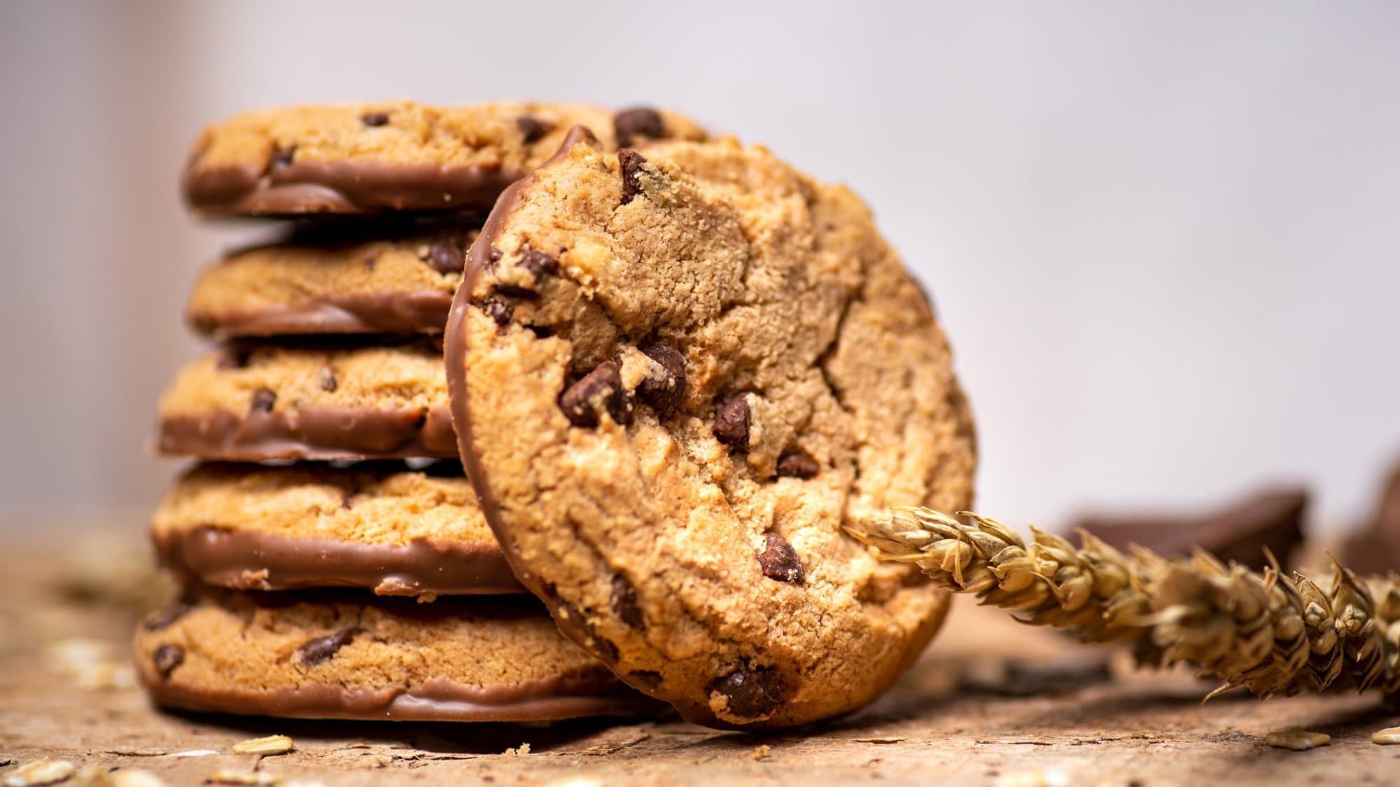 Frontalsicht: An einem Stapel Peanut and Chocolate Cookies, ist ein weiterer Keks angelehnt. Daneben liegt eine Weizenähre.