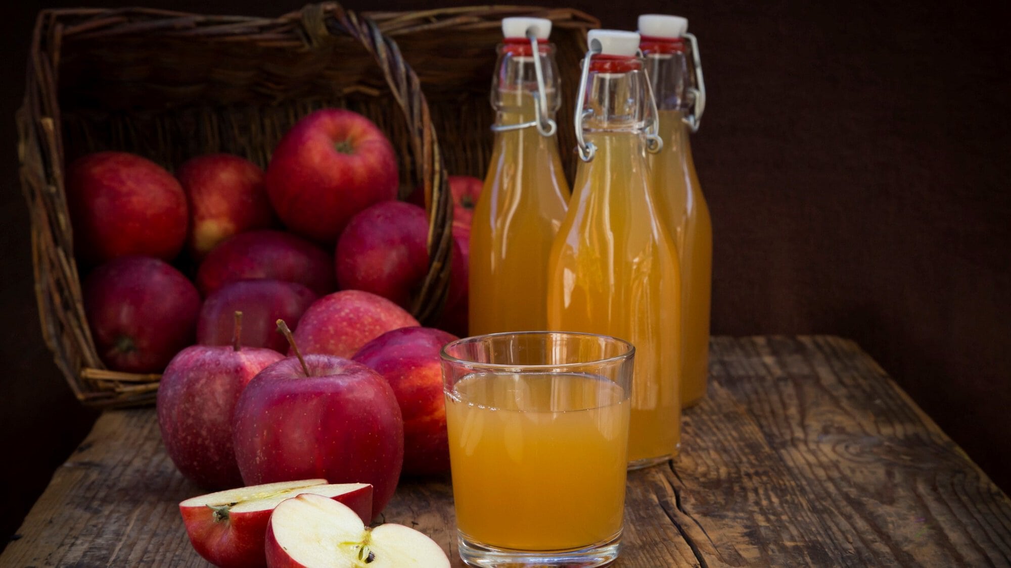 Frontalsicht: Drei Flaschen sekbst gemachten Apfelsaft, sowie mehrere Äpfel und Apfelhälften stehen auf einem Holztisch vor einem dunklen Hintergrund.