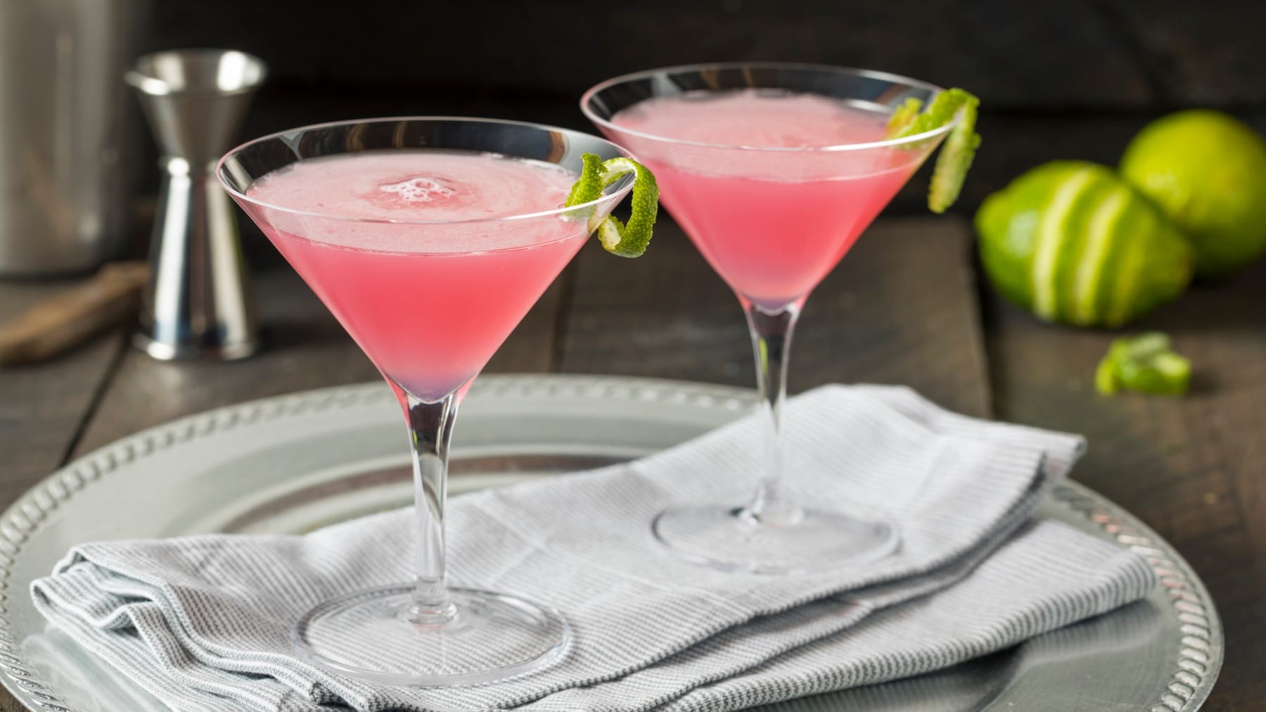 Zwei Martinigläser Pink Cosmo garniert mit Limettenschale auf einem hellen Tablett vor dunklem Hintergrund.