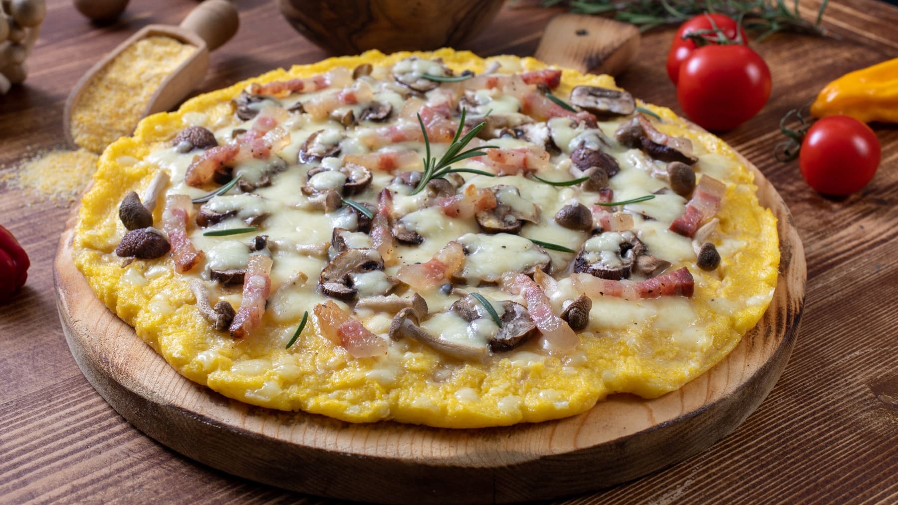 Die Polenta-Pizza auf einem runden Holzschneidebrett. Daneben Maisgrieß, Kirschtomaten und Pilze.