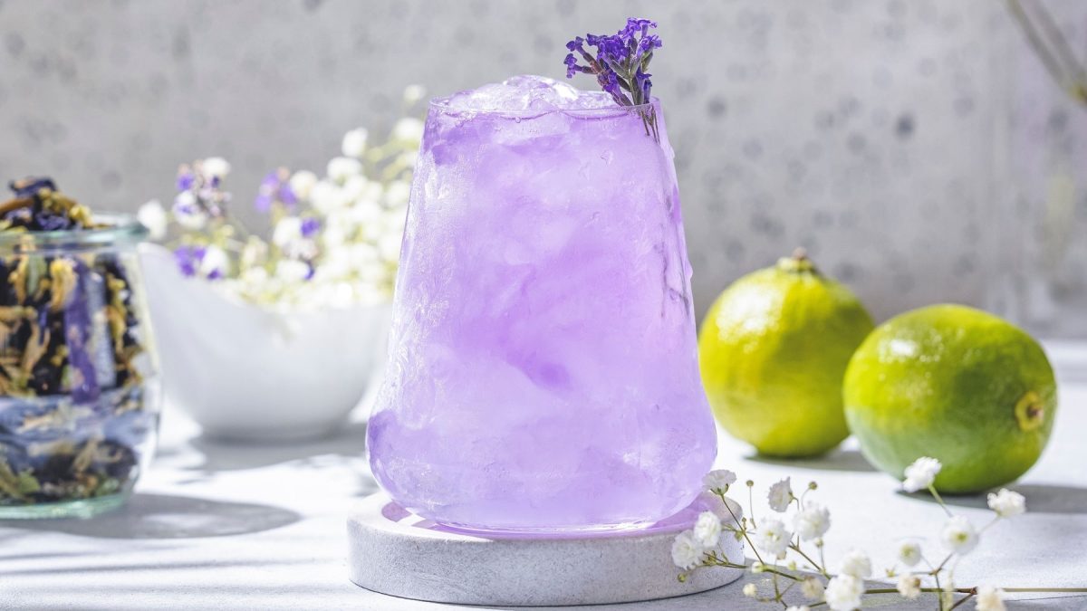 Ein Glas mit dem violetten Cocktail auf hellem Untergrund. Im Hintergrund Limetten, Anchan-Tee und lila Blüten.
