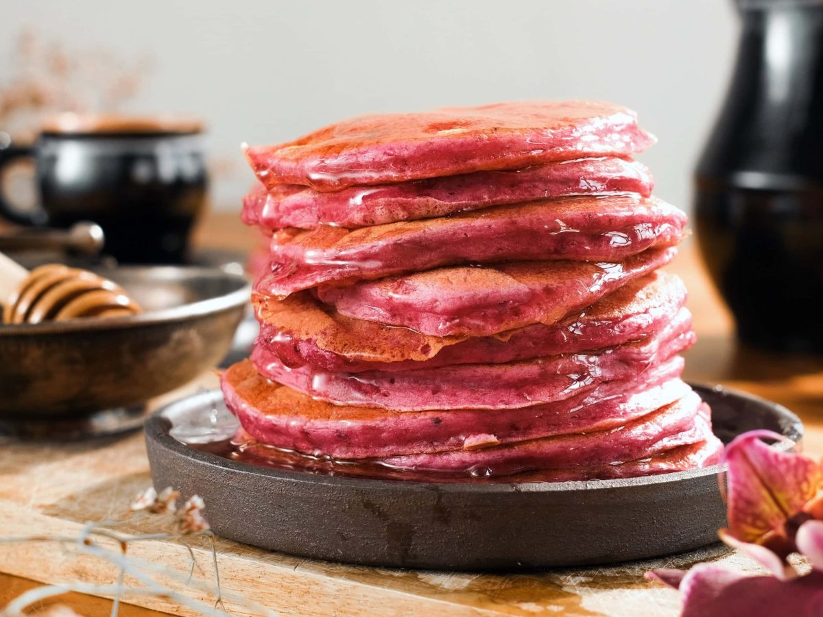 Ein schwarzer Teller mit den gestapelten Rote Bete Pancakes auf einem Holztisch mit viel schwarzem Geschirr im Hintergrund.