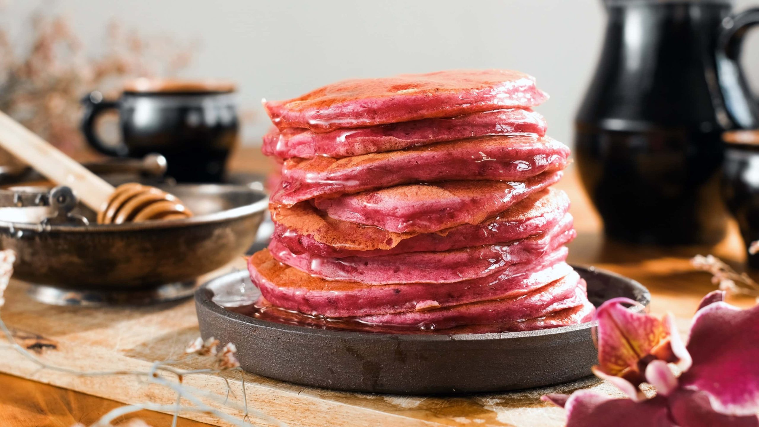 Ein schwarzer Teller mit den gestapelten Rote Bete Pancakes auf einem Holztisch mit viel schwarzem Geschirr im Hintergrund.