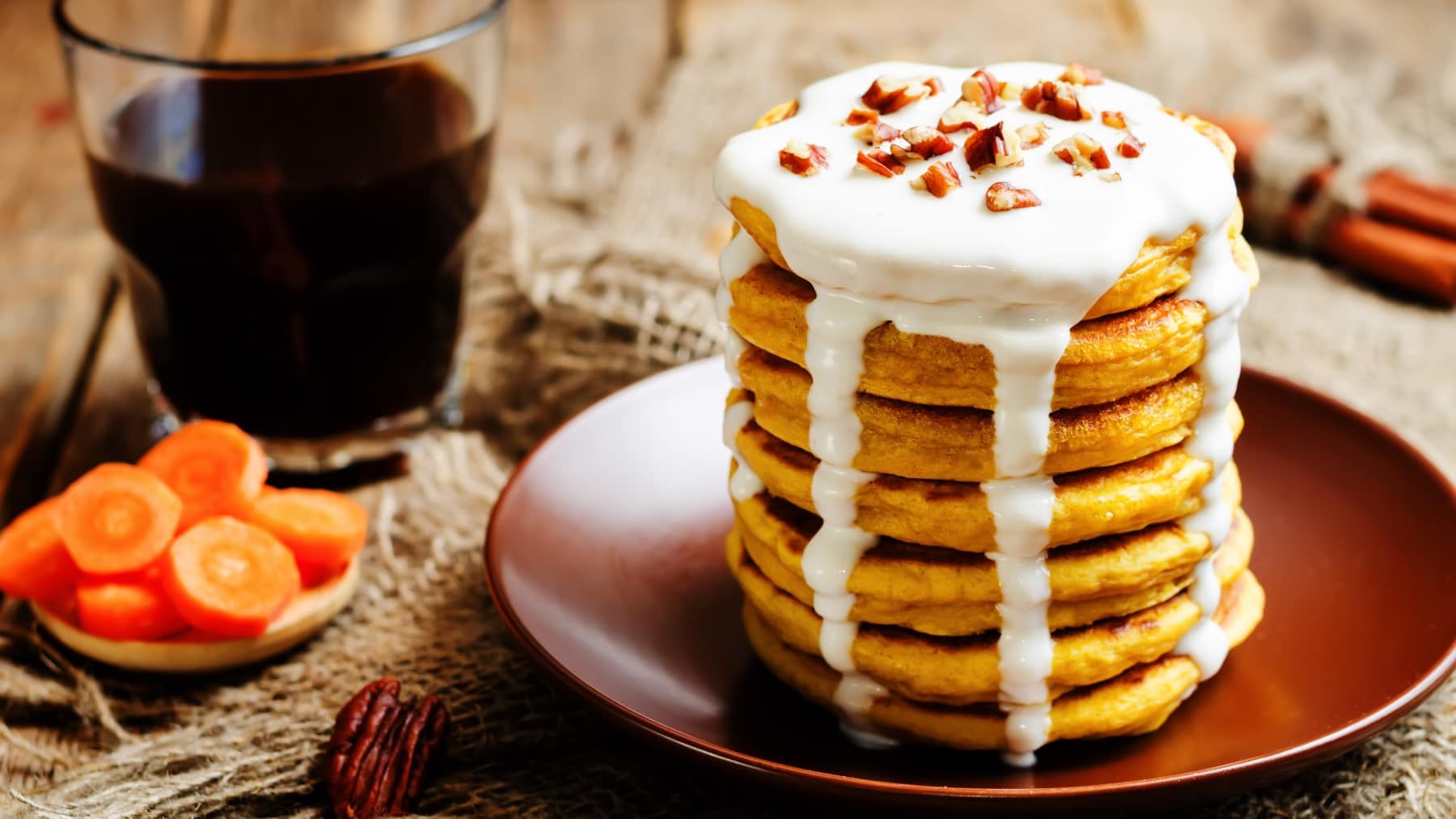 Ein Stapel Rübli-Pancakes mit Frosting-Sauce und gehackten Nüssen auf einem braunem Teller neben einem Glas Kaffee und Möhrenscheiben.
