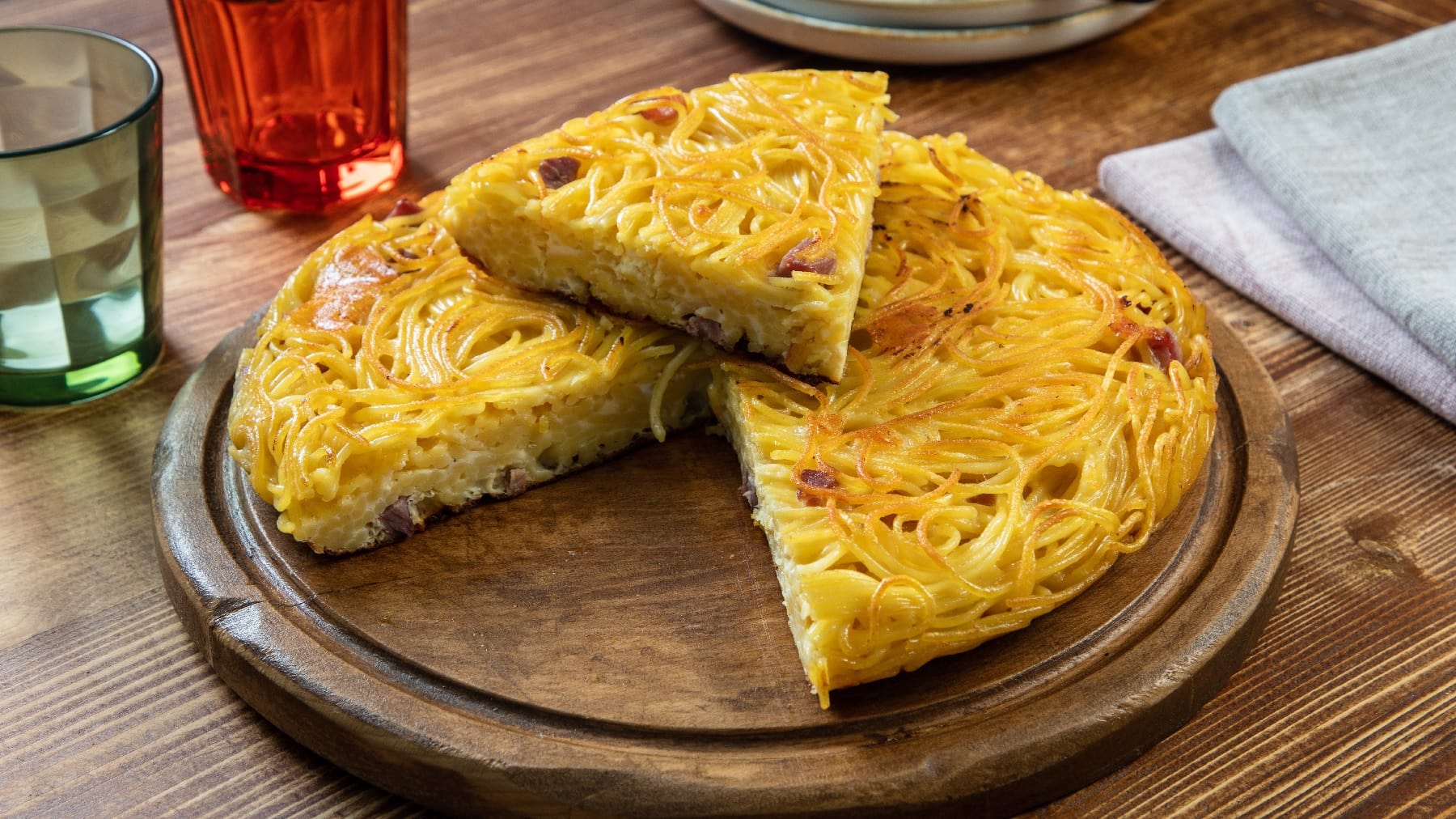 Der Spaghetti-Carbonara-Kuchen auf einem Holzbrett und einem Holztisch. Im Hintergrund unscharf zwei Gläser.