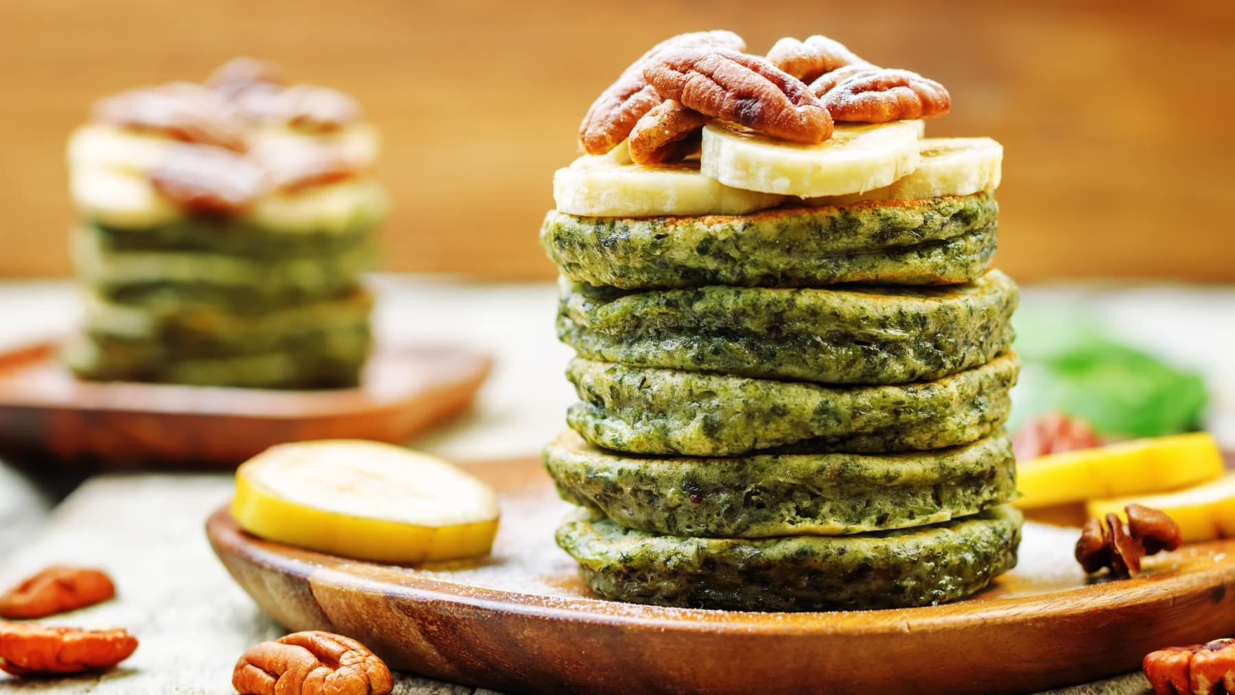 Gestapelte Spinat-Pancakes mit Bananenscheiben und Pekannüssen darauf auf einem Holzteller.