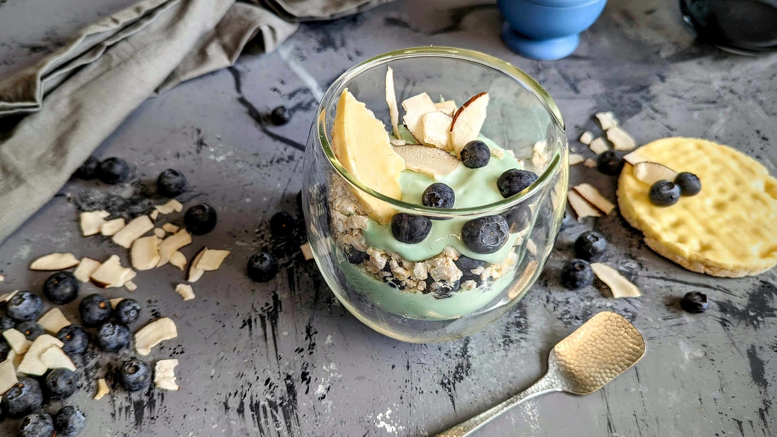 Ein Glas mit dem geschichteten Frühstück aus Spirulina-Joghurt, Blaubeeren , Resiwaffel und Kokoschips auf einem grauen Marmoruntergrund.