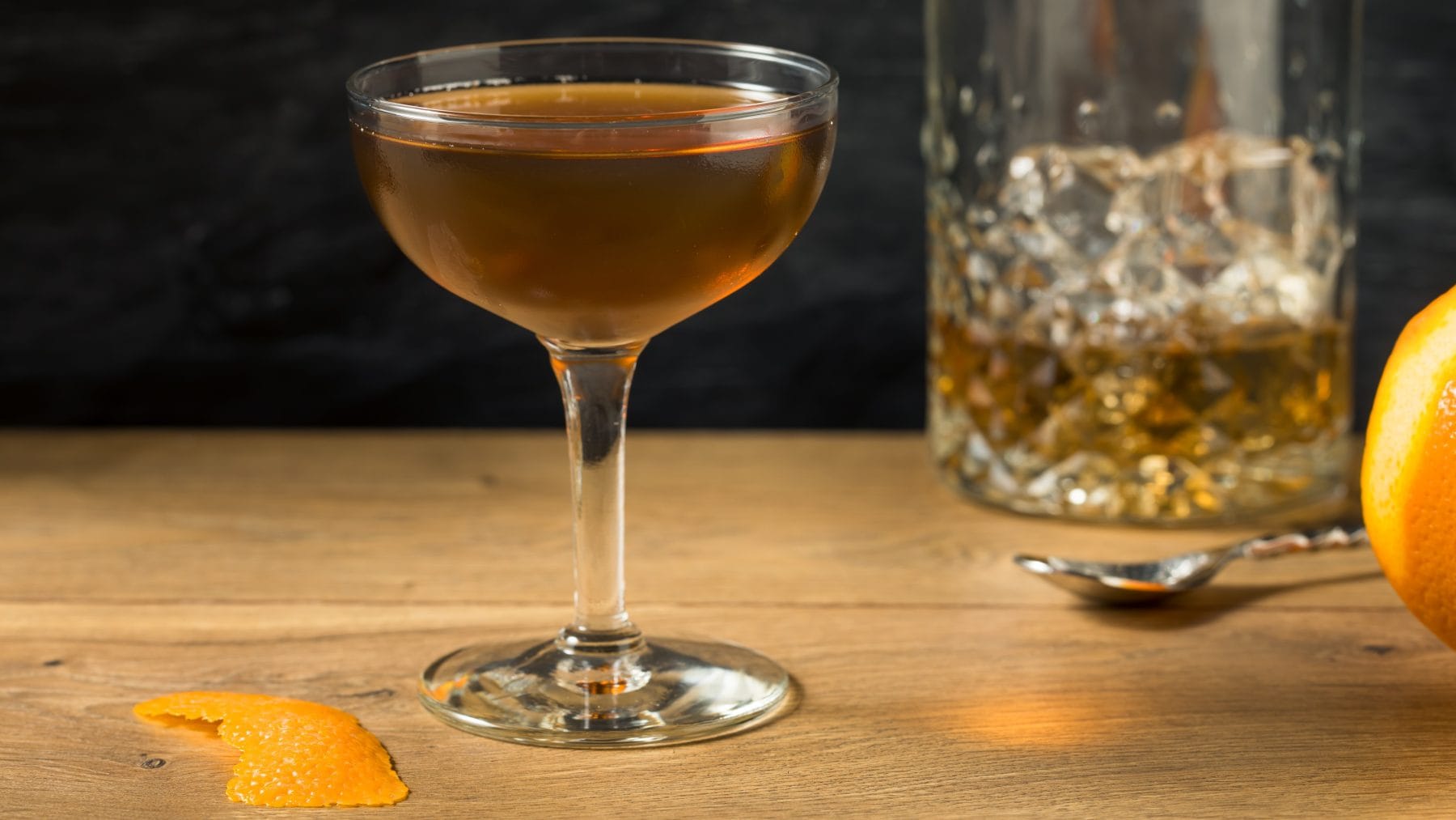 Tipperary Cocktail No.1 mit Proper No. Twelve im Glas auf Holztisch. Im Bild ein Whiskey Glas, Orange und Orangenzeste.