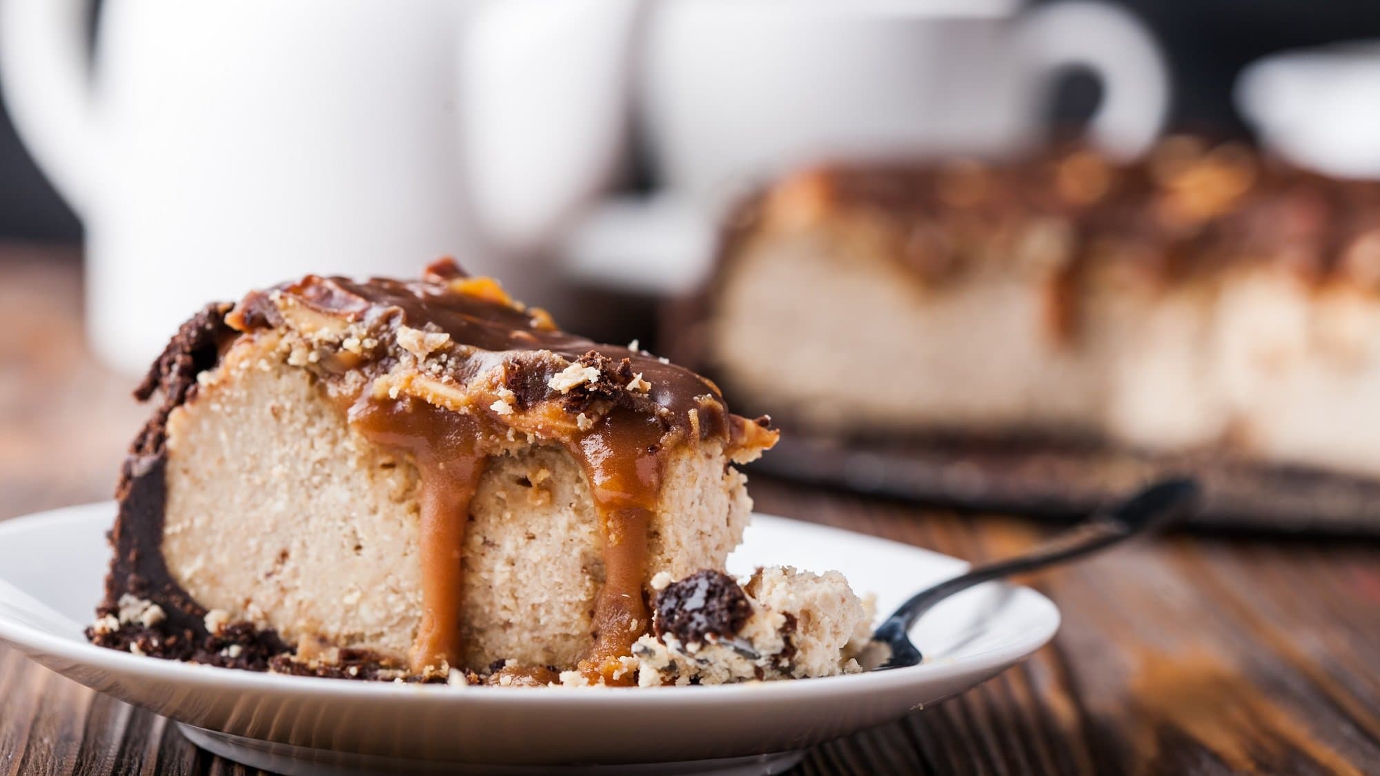 Ein Stück Erdnussbutter-Cheesecake mit Schokosauce und gehackten Erdnüssen garniert auf einem Teller. Im Hintergrund unscharf der restliche Kuchen.