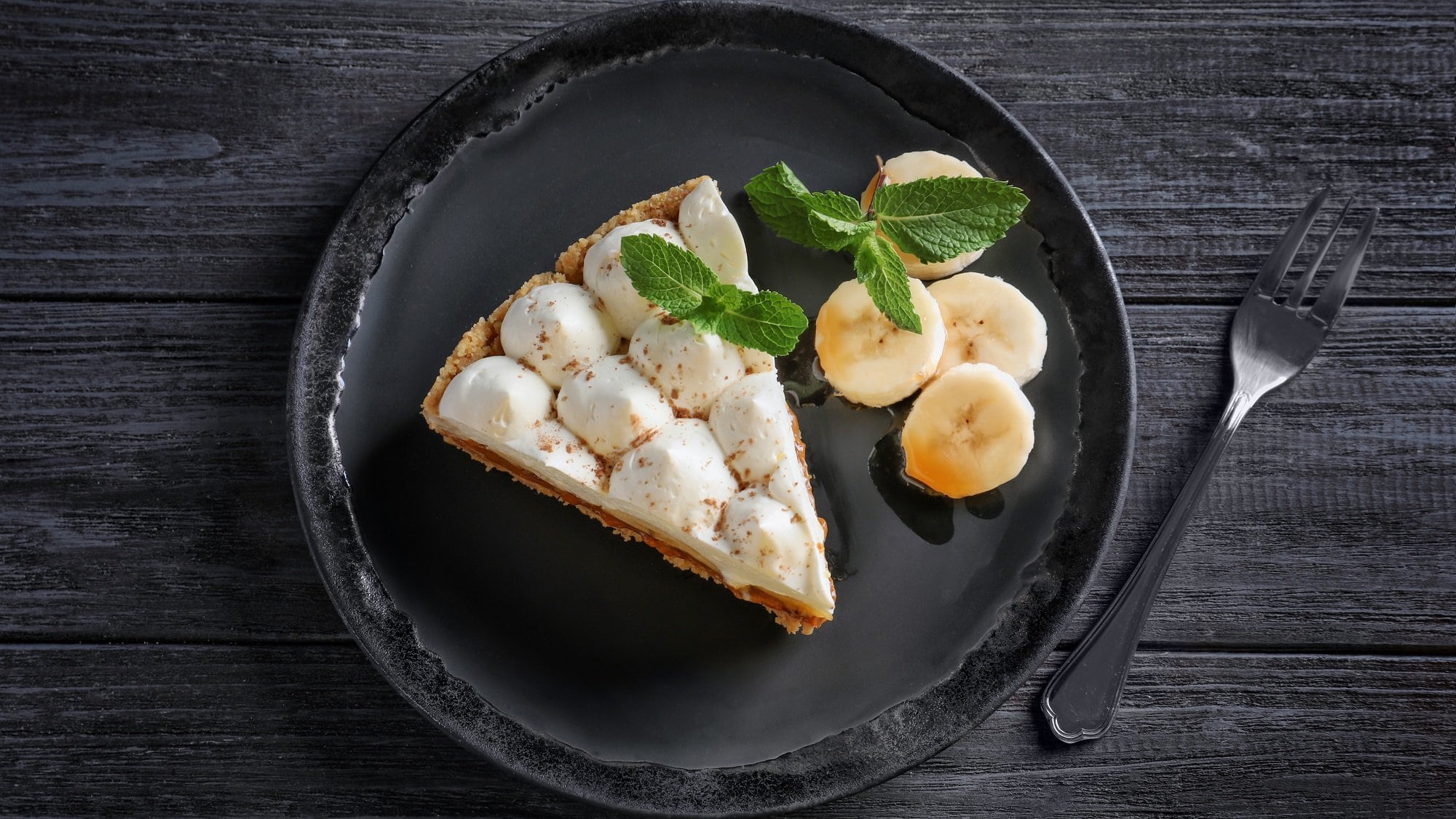 Ein Stück veganer Karamell-Kuchen mit Kokoscreme auf einem Teller zusammen mit Bananenscheiben, daneben eine Kuchengabel. Draufsicht.