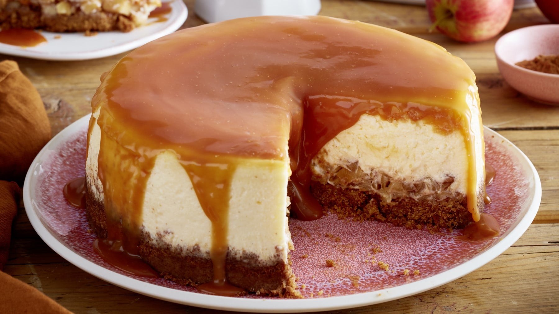 Angeschnittener Apfel-Karamell-Cheesecake auf einem rosa Teller mit Holz und Geschirr im Hintergrund.